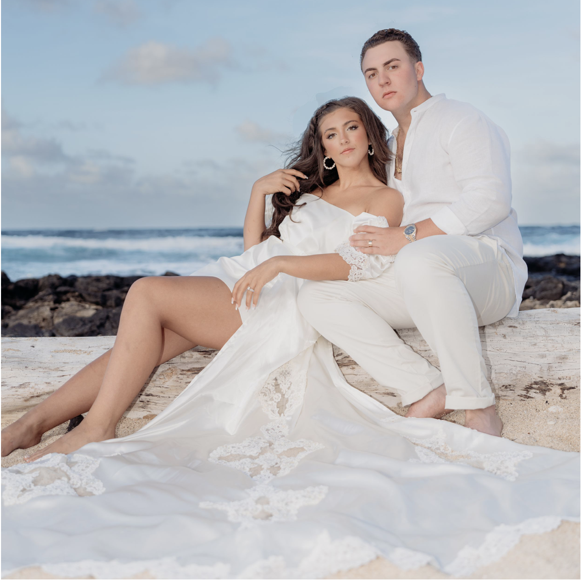 A young couple in white clothing sitting on a beach with the ocean and cloudy sky in the background, with the woman wearing a white dress and the man in a white shirt and pants.