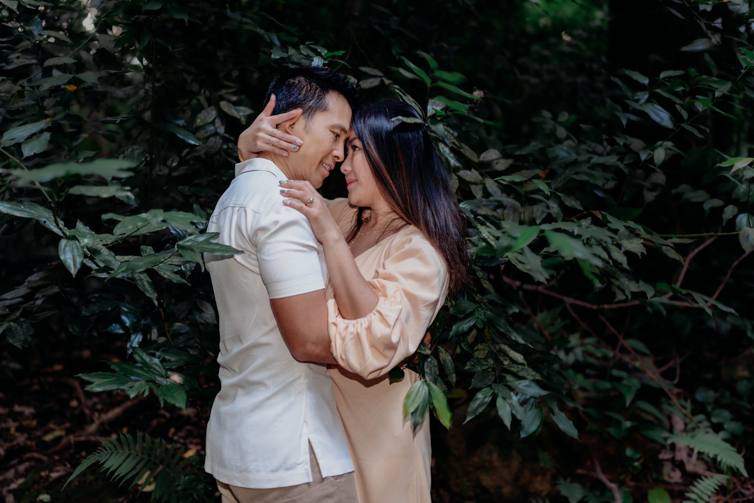 A couple in white and beige clothes embrace and hold each other closely in a forested area, touching foreheads and smiling.