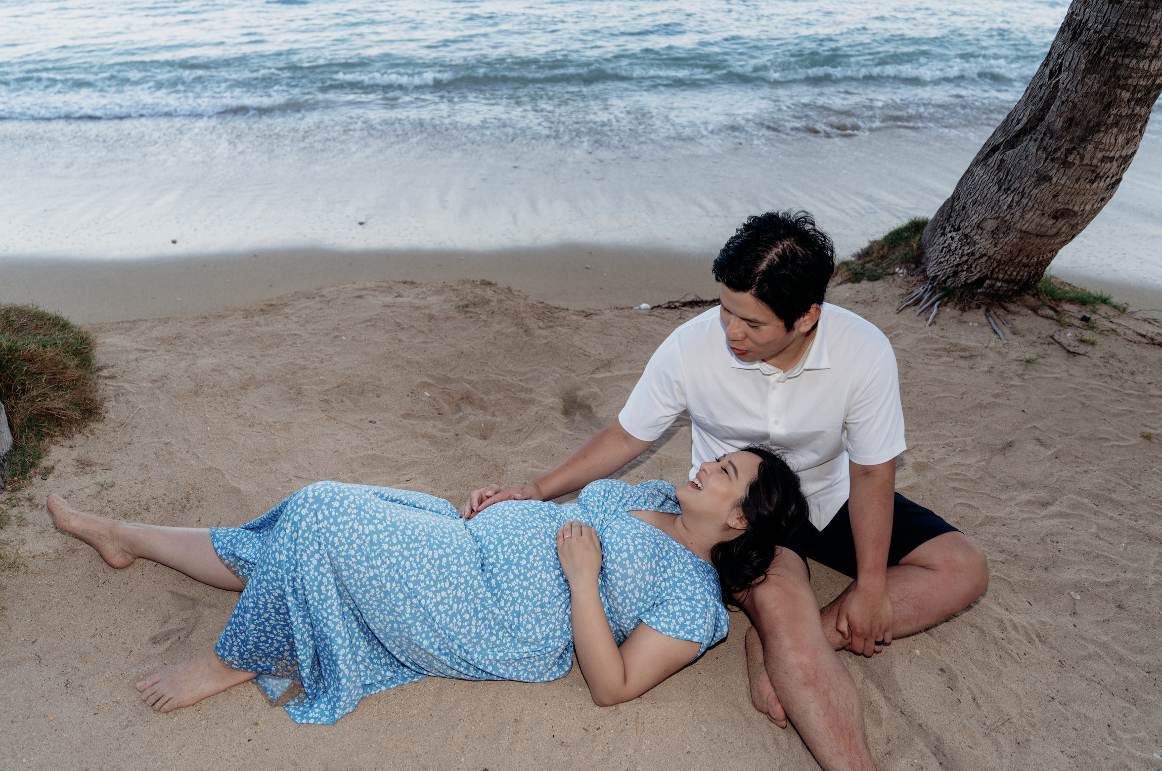 A man and woman on a beach, with the woman lying on her side on the sand and pregnant, wearing a blue dress with white floral pattern, and the man sitting beside her, holding her hand and looking at her. There is a tree leaning over the sand near the