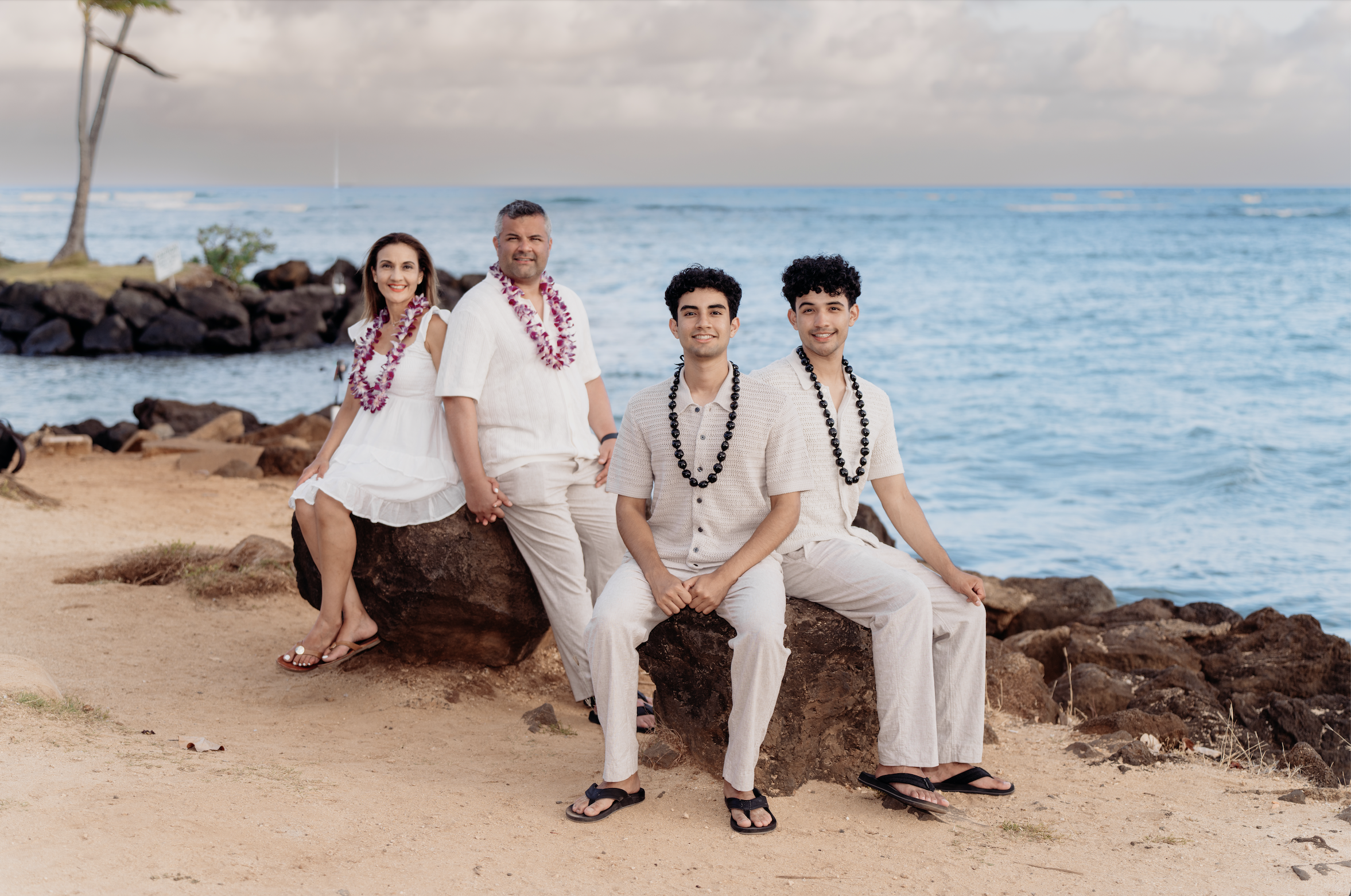 A family of five dressed in white and wearing leis, sitting and standing on rocks at the beach with ocean and cloudy sky in the background.
