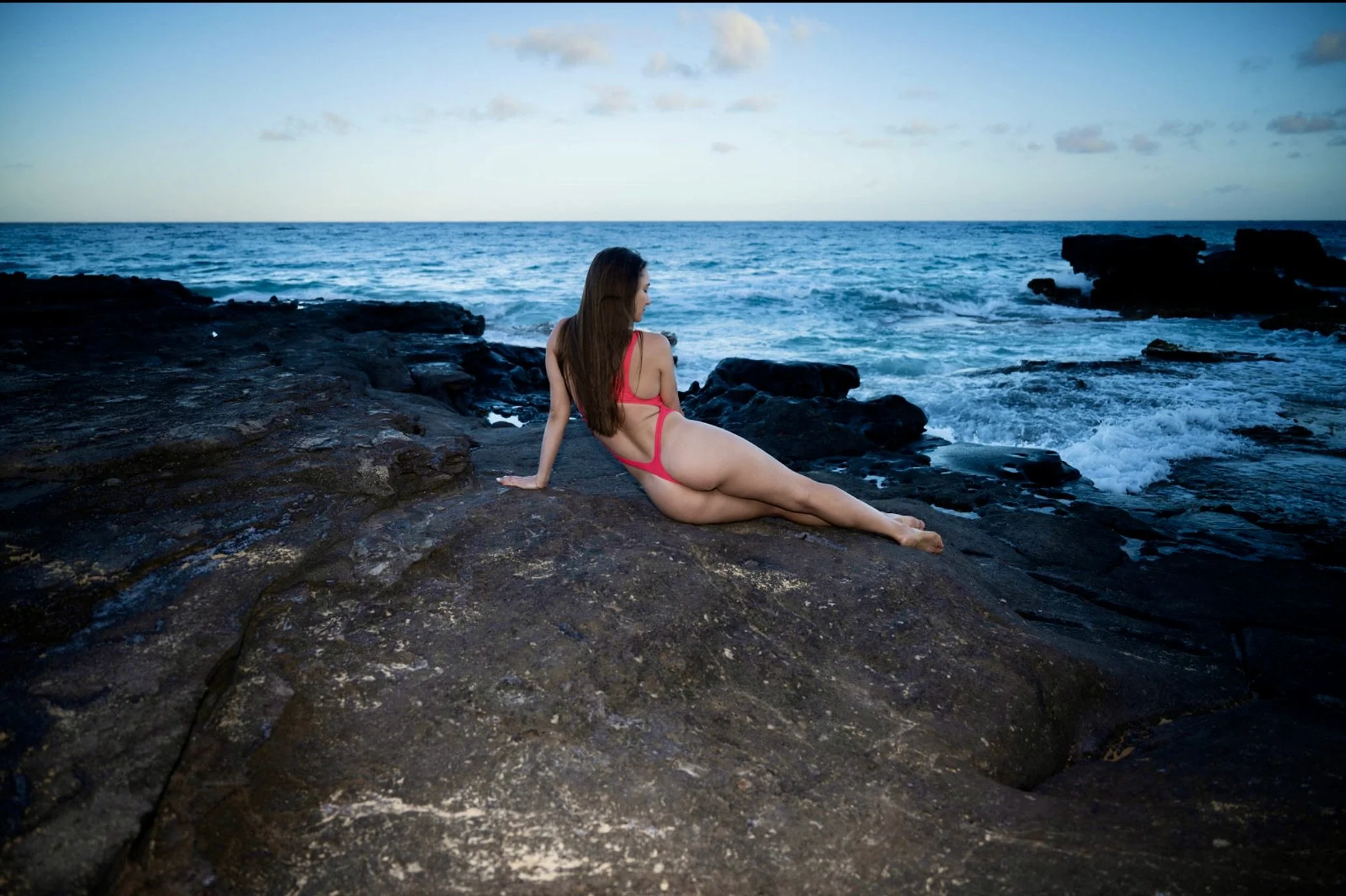 A woman in a pink swimsuit sitting on rocks by the ocean at sunset, looking at the water.