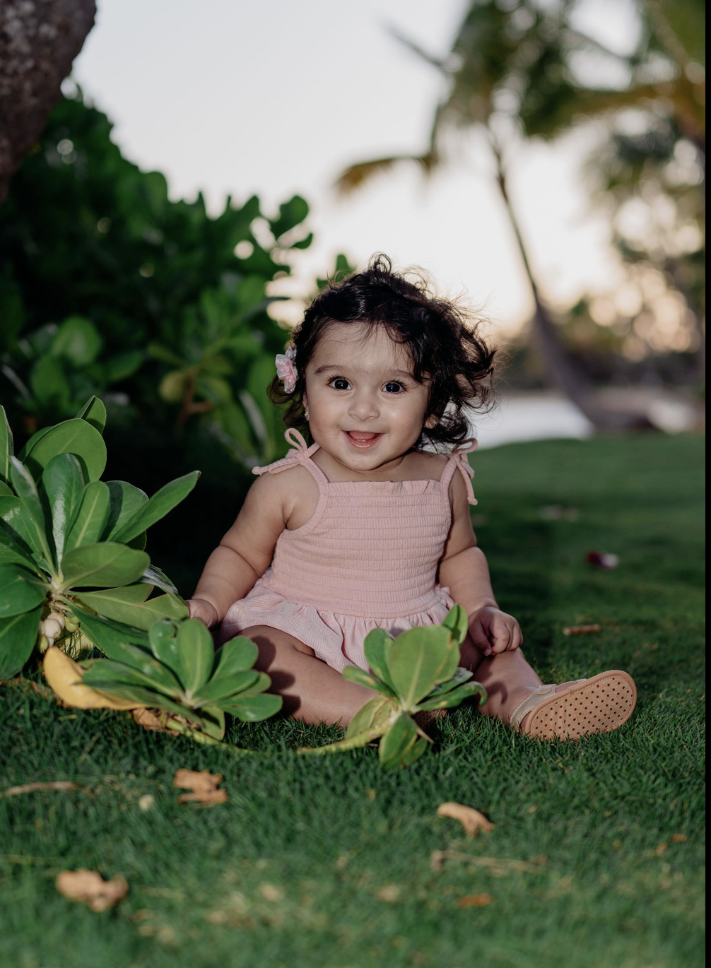 A smiling baby girl with curly dark hair, wearing a pink dress, sitting on green grass near tropical plants, outdoors during sunset.