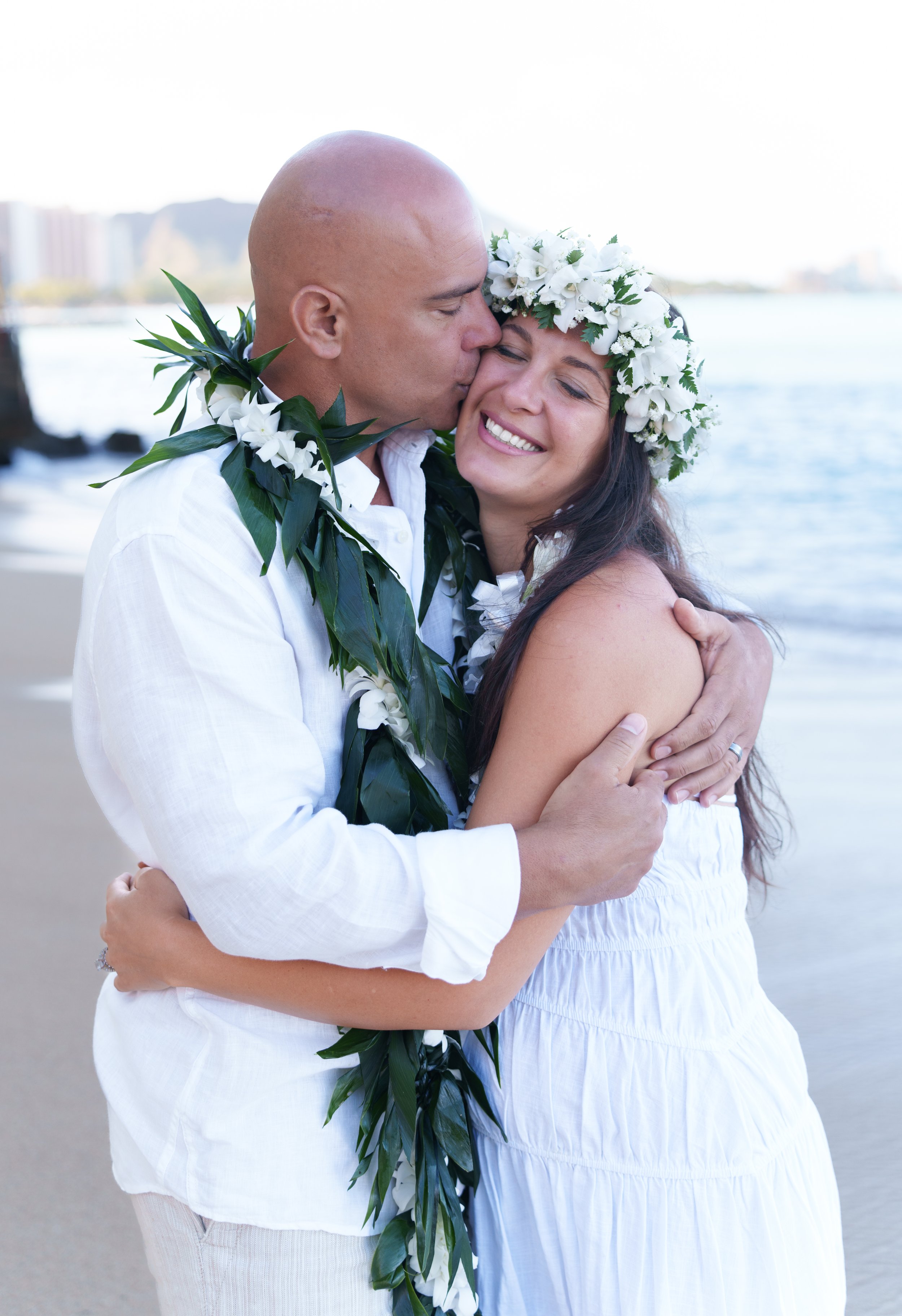 A couple in white clothing and floral leis sharing a hug and a kiss by the water, both wearing white floral crowns, during a beach wedding or celebration.
