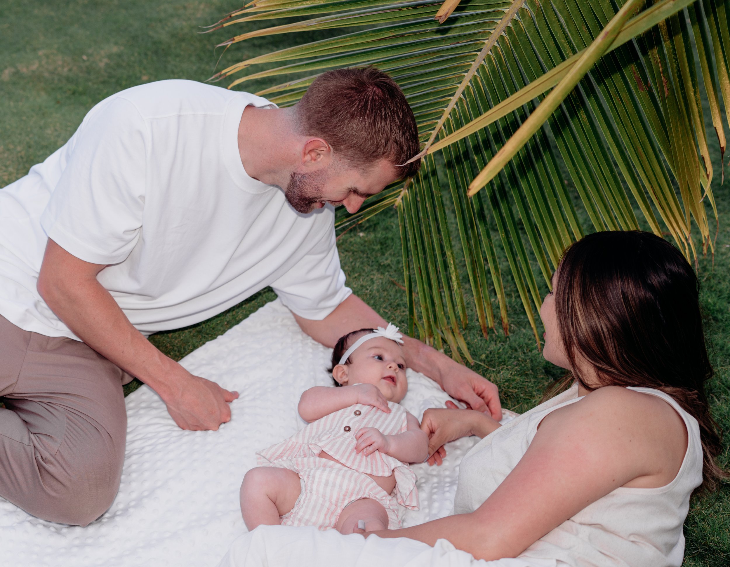 A man and woman with a baby girl lying on a white blanket outdoors under palm tree, with grass in the background.