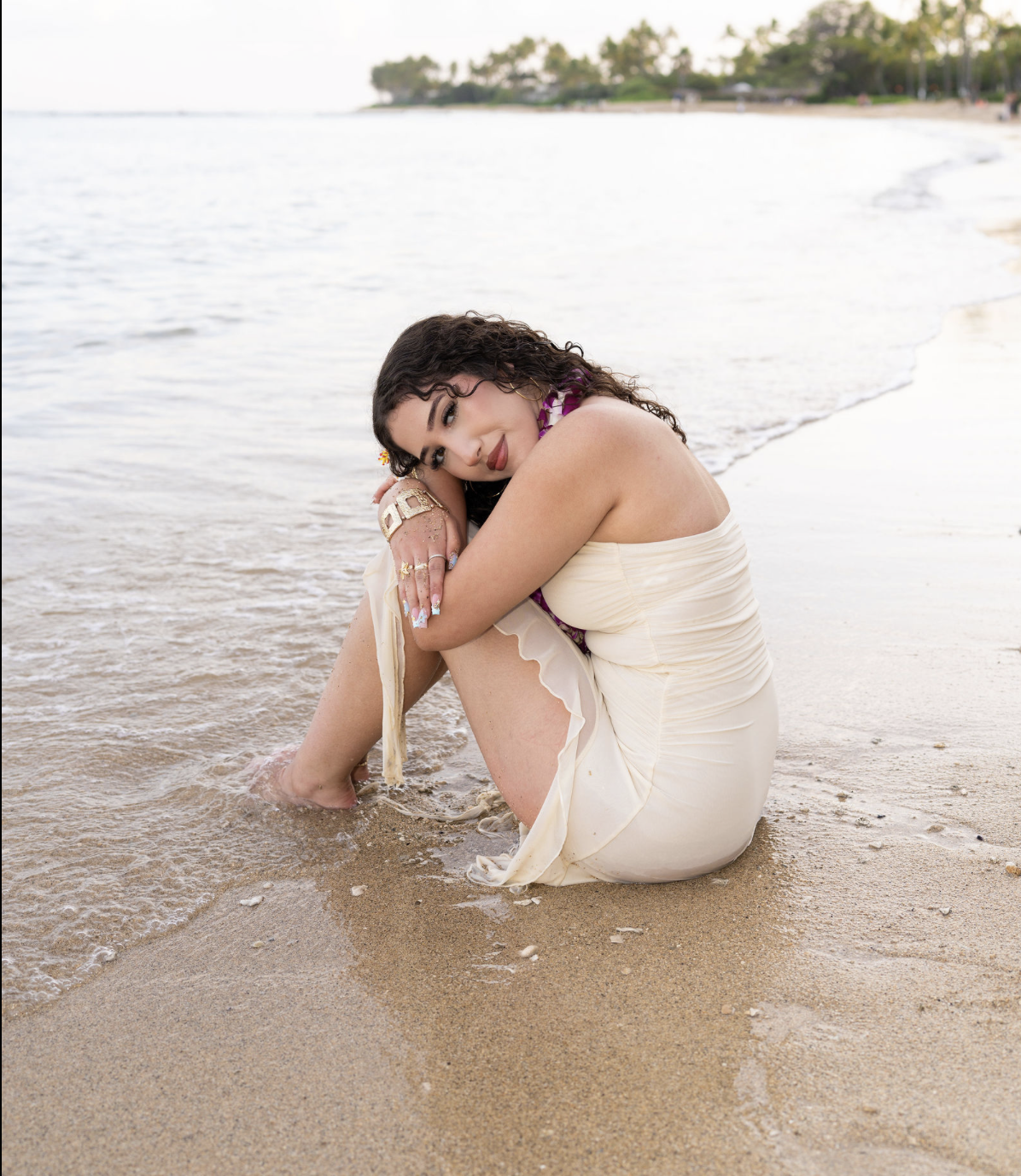 A woman sitting on the sandy beach near the water, with her arms wrapped around her knees, looking at the camera.