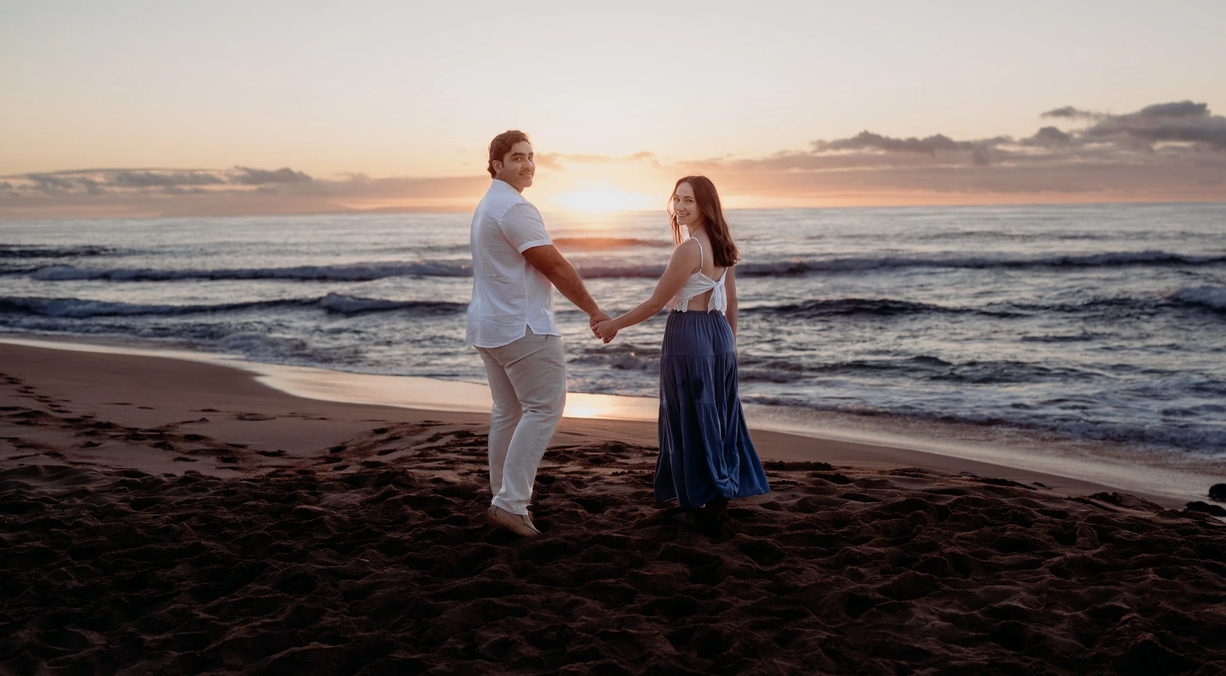 A couple holding hands on the beach during sunset, with the ocean behind them.