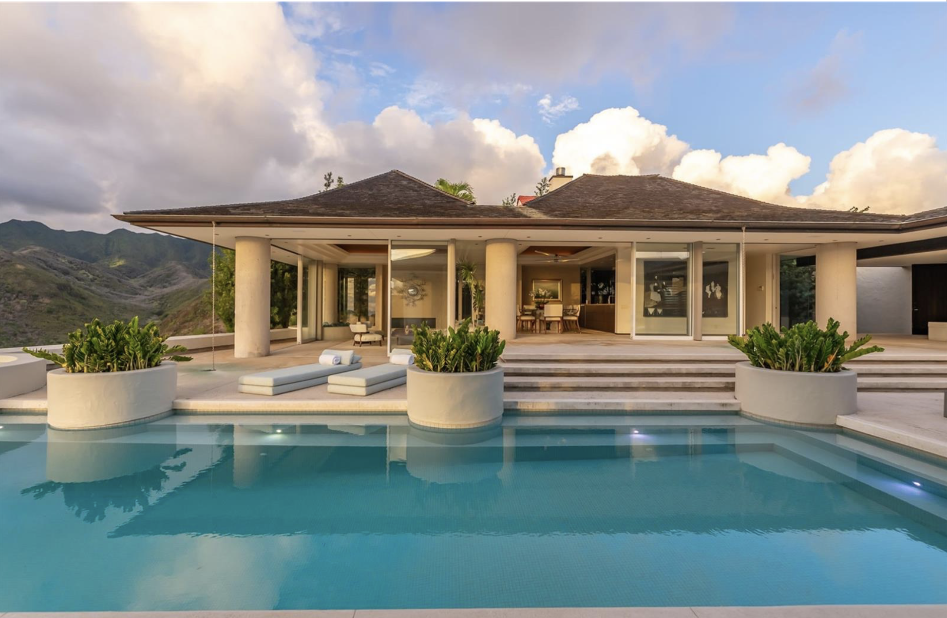 Luxury house with a swimming pool in front, surrounded by potted plants, outdoor lounge chairs, and a mountainous landscape in the background.
