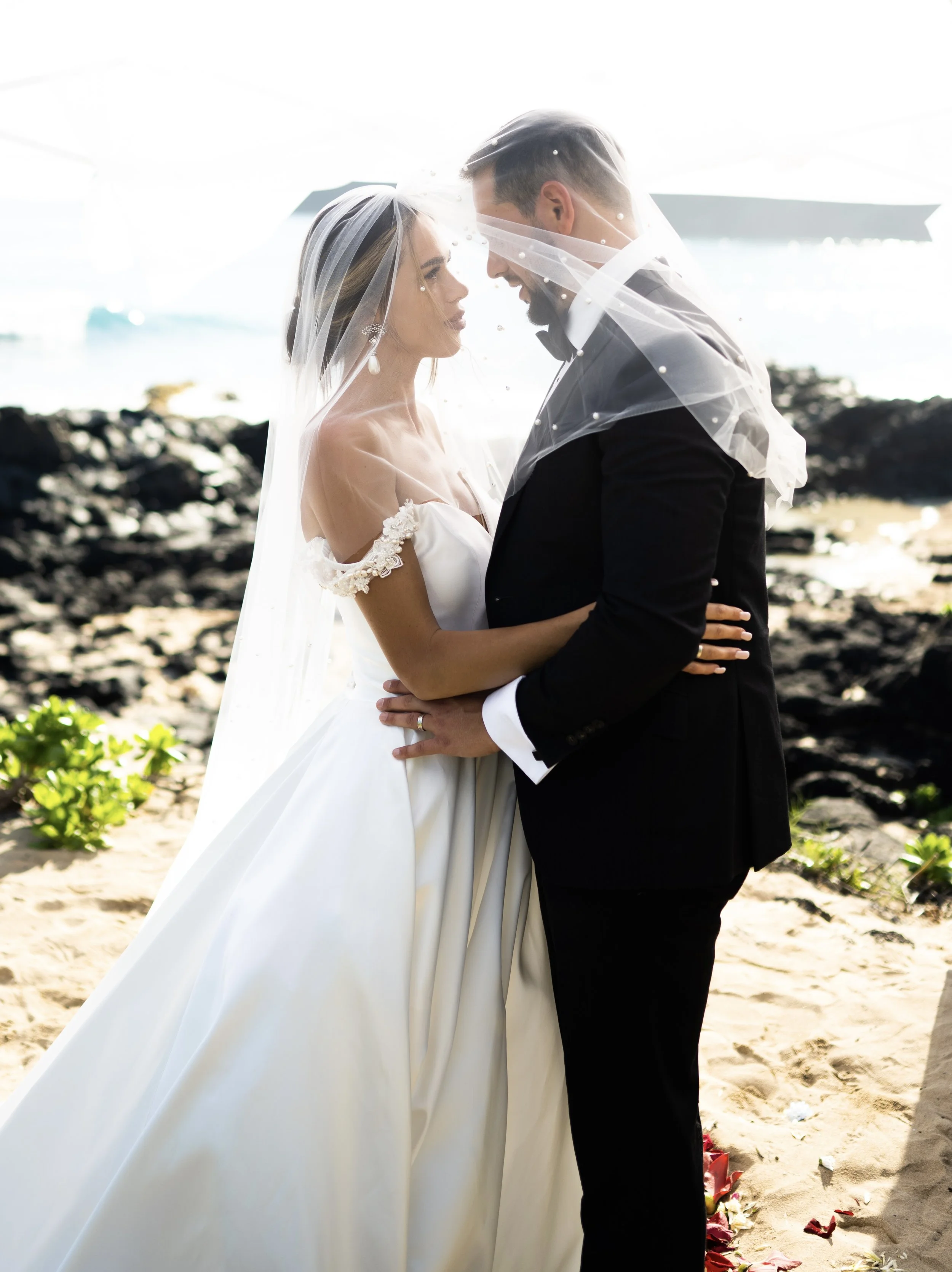A bride and groom standing close on a beach, facing each other, with the bride wearing a white gown and veil, and the groom in a black tuxedo and veil, embracing each other under an umbrella.