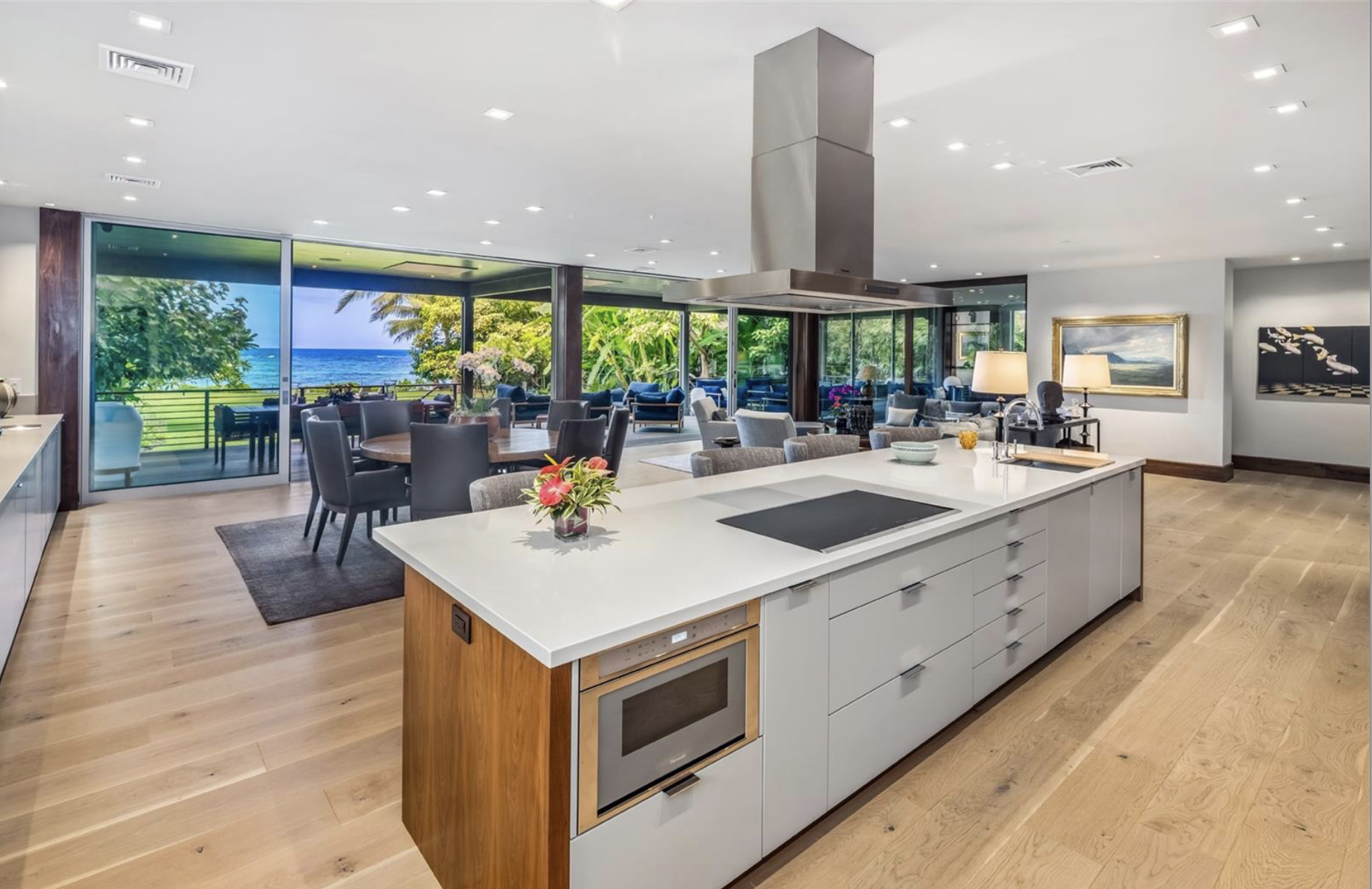Open-concept living space with a view of the ocean through large glass windows, including a kitchen island with a stove, dining area, and sitting area with outdoor view.