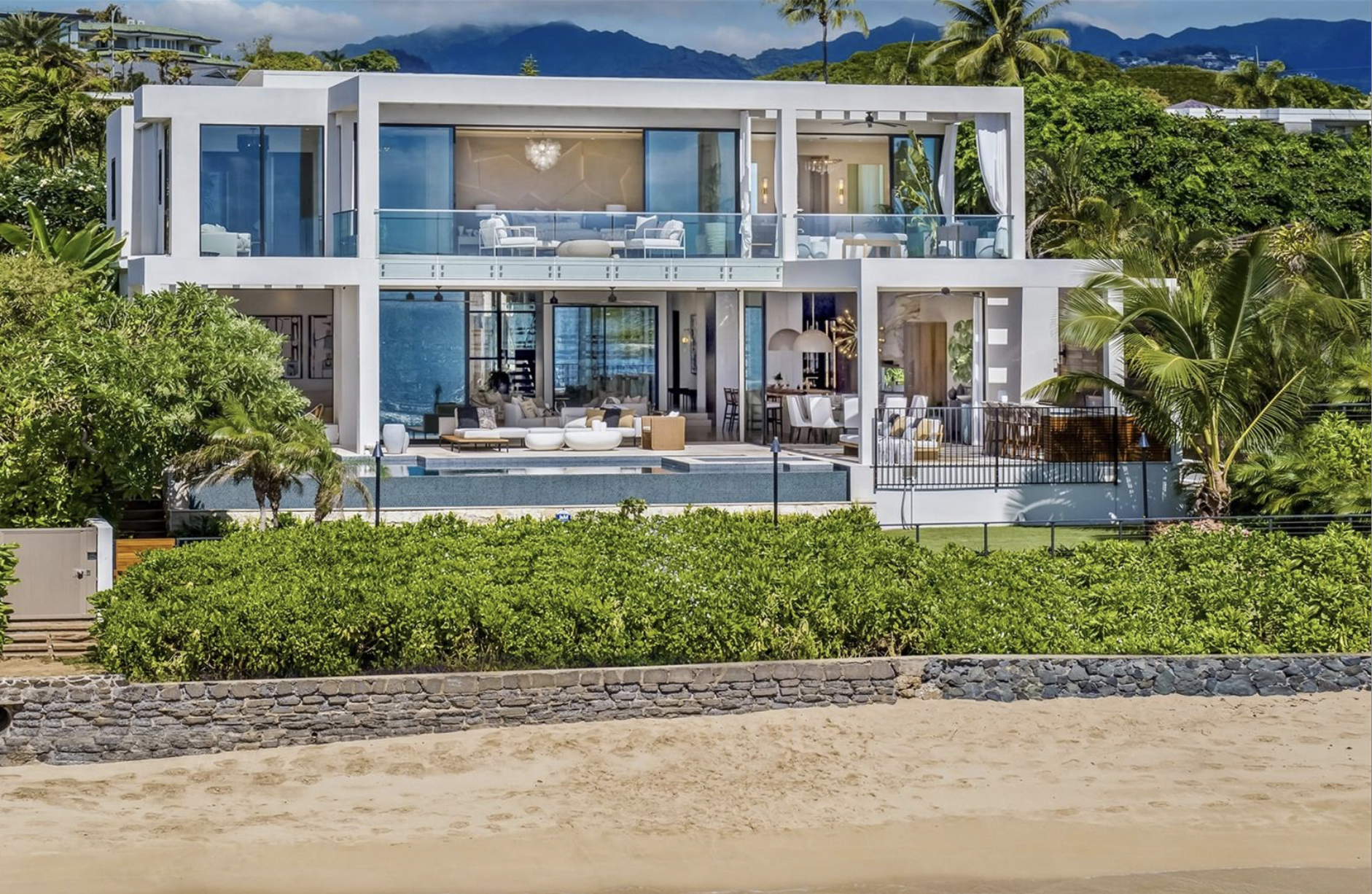 Modern beachfront house with multiple outdoor seating areas and a swimming pool, surrounded by lush green foliage and palm trees, with mountains in the background.