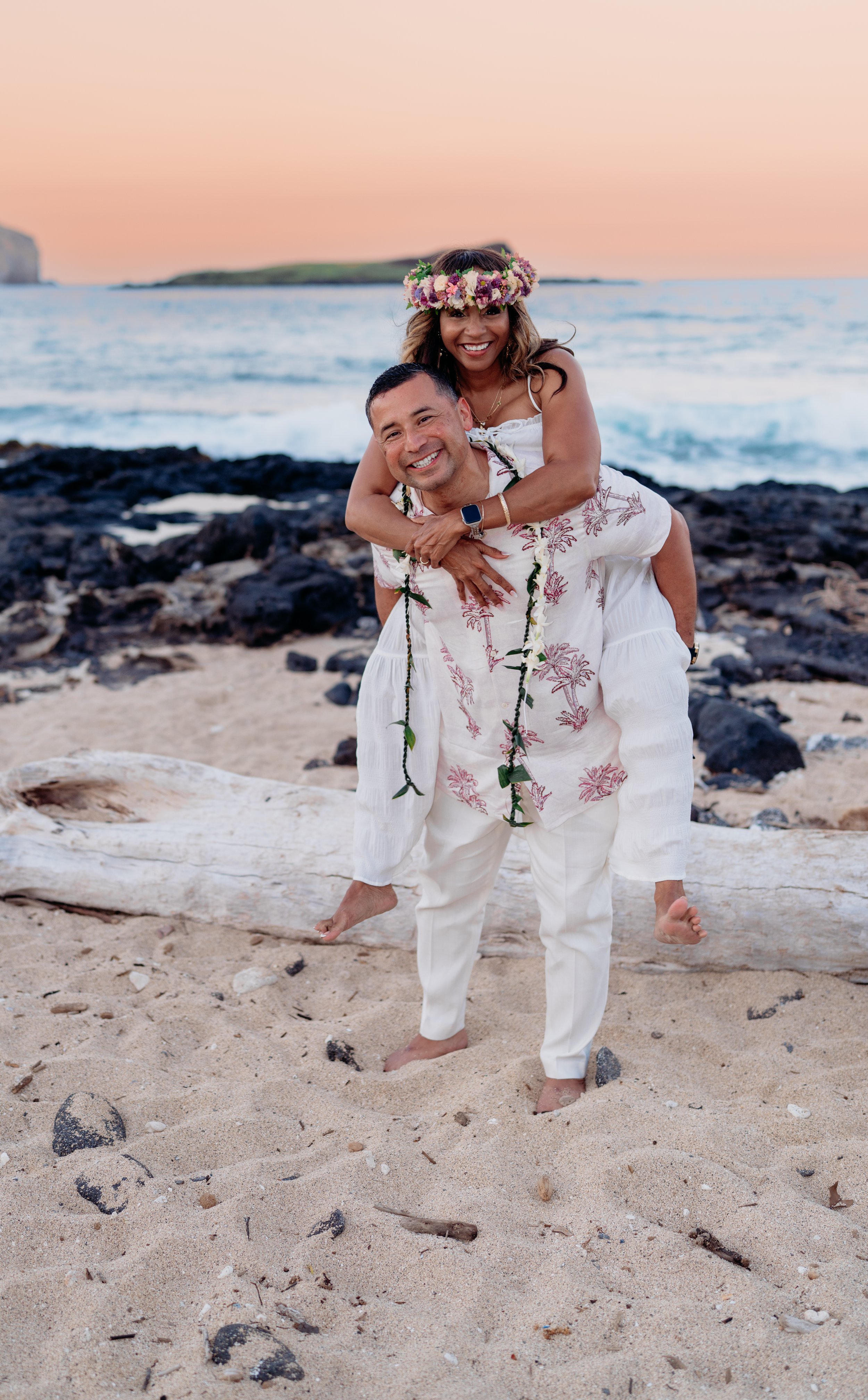 A happy couple on a beach at sunset, with the woman giving the man a piggyback ride. The woman wears a flower crown and a white dress, while the man wears a floral shirt and white pants. They are smiling and looking at the camera, with ocean waves an