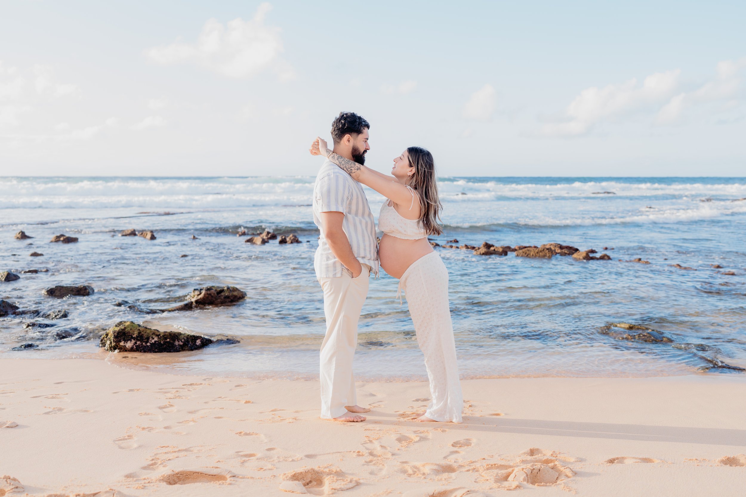 Pregnant woman and man standing on the beach near the water, embracing and looking at each other.