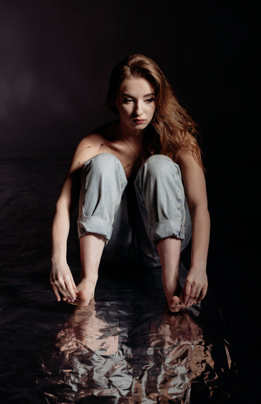 A woman with red hair crouching on a reflective surface in front of a dark background.