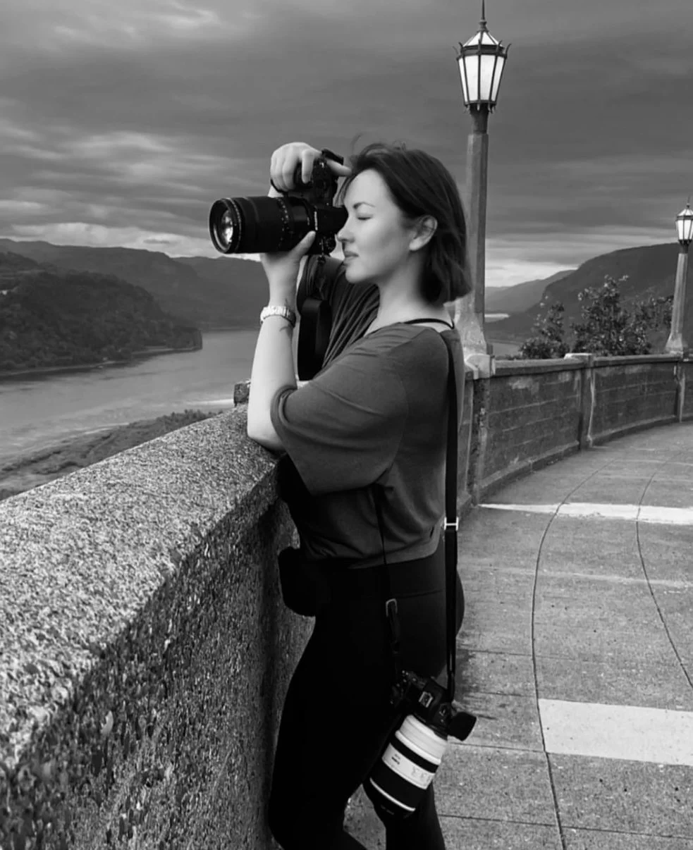 A woman in profile, standing on a bridge, taking a photograph with a camera. She has shoulder-length hair, is dressed casually, and has a camera with a zoom lens hanging from her neck. The scene behind her features a river, hills, and a cloudy sky, and there are vintage-style street lamps along the bridge.