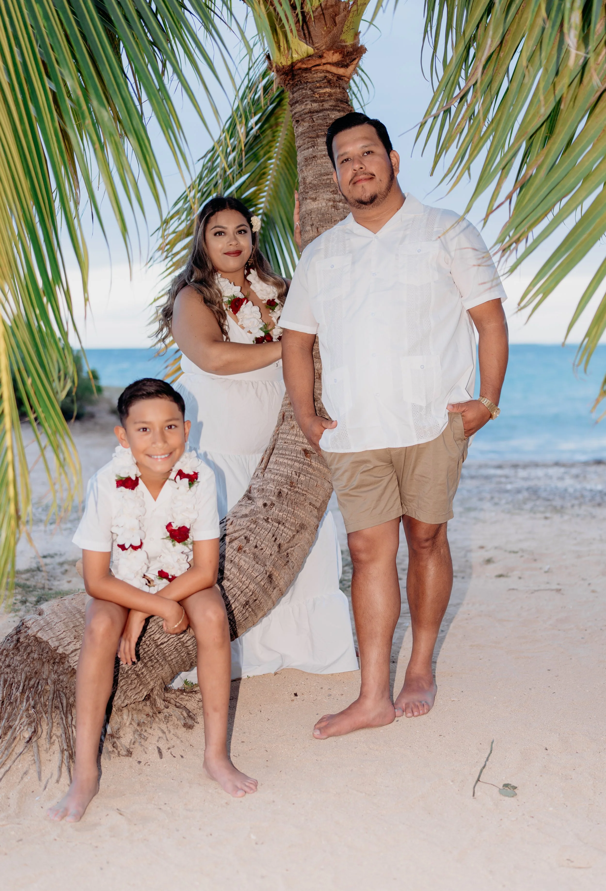 A family of three posing on a beach with a palm tree and ocean in the background. They are dressed in white clothing and wearing flower leis, smiling at the camera.
