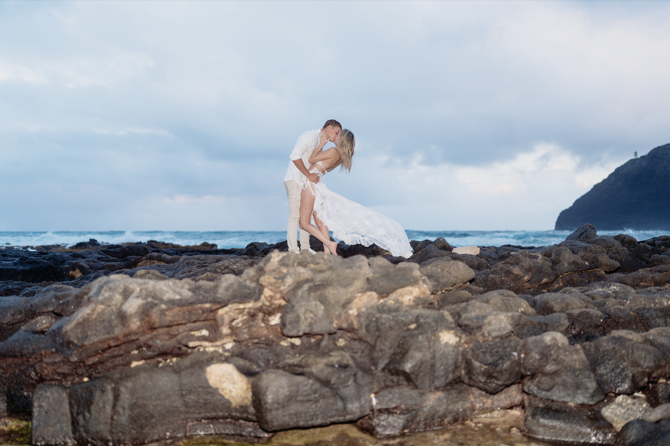 Couple in wedding attire kissing on rocky beach with ocean and mountainous landscape in background.