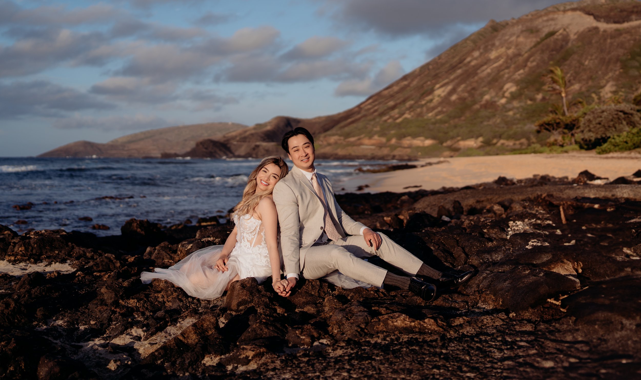 A couple dressed in wedding attire sitting on rocks by the ocean in a scenic coastal landscape with mountains in the background.