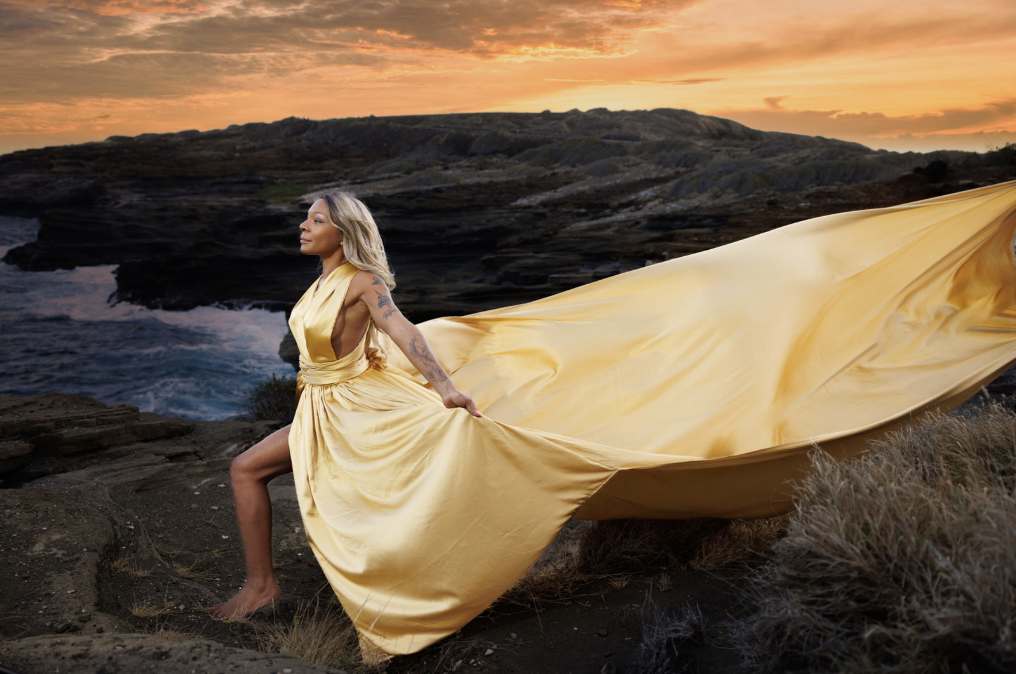 Woman in a flowing yellow dress standing on rocky terrain by the ocean during sunset.