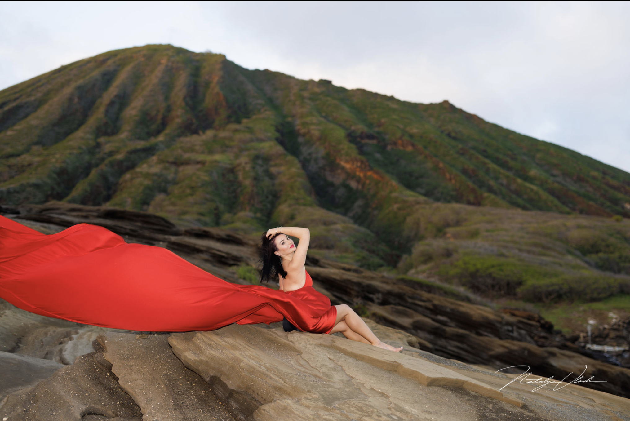 Woman in a red dress sitting on rocks with a mountain background.