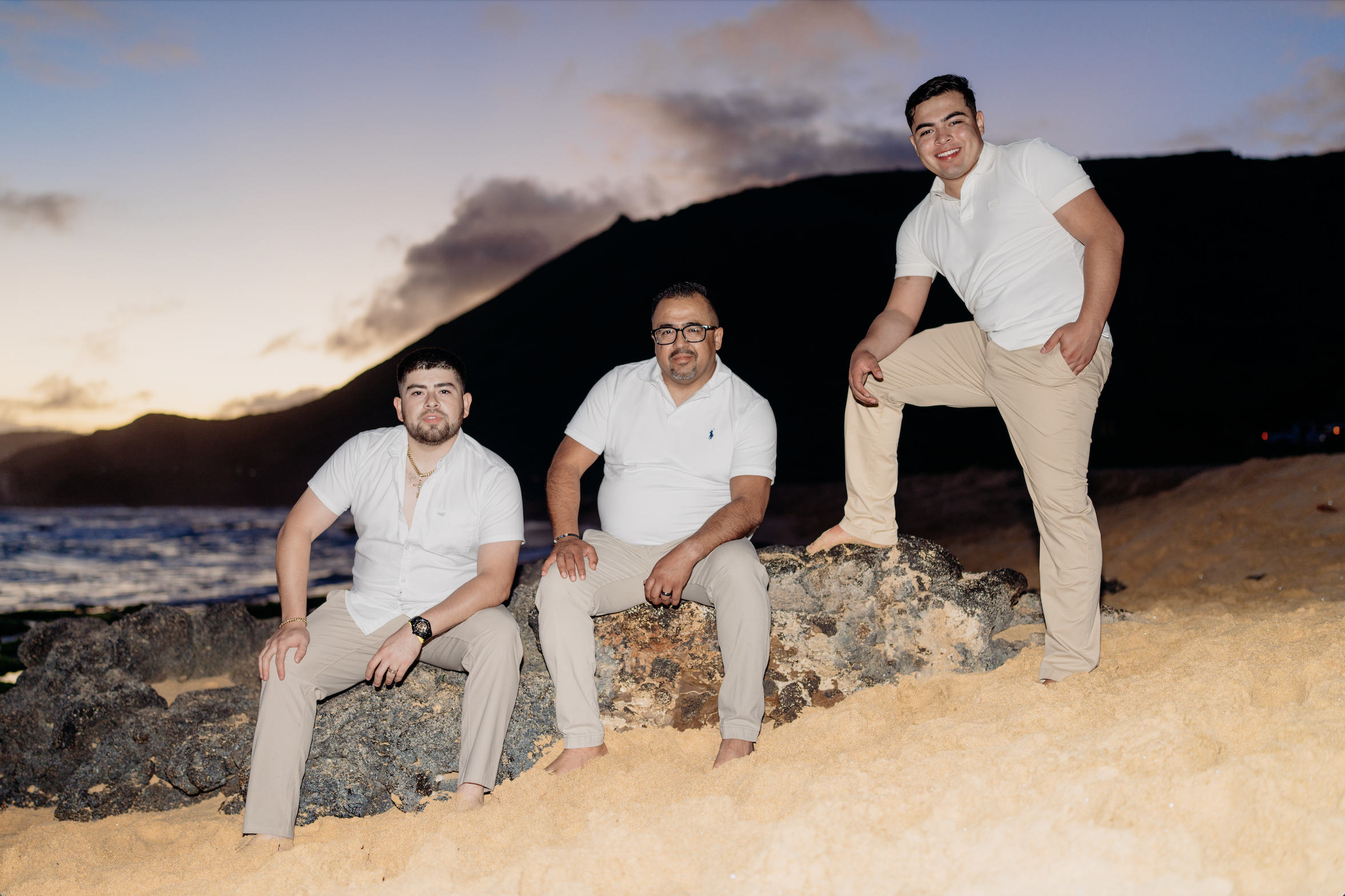 Three men on a beach at sunset, sitting and standing on rocks, with a mountain and the ocean in the background, wearing white shirts and beige pants, smiling.