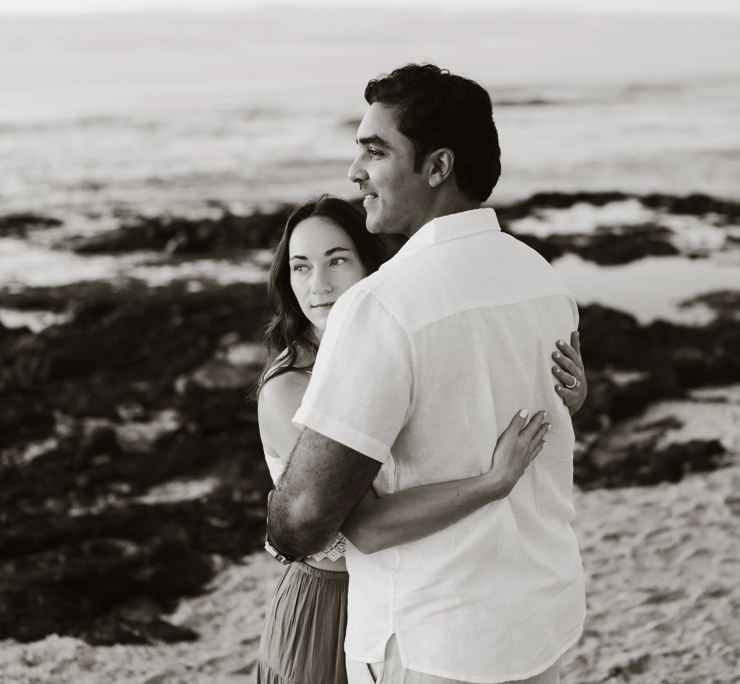 A couple embracing on a beach with rocks and water in the background.