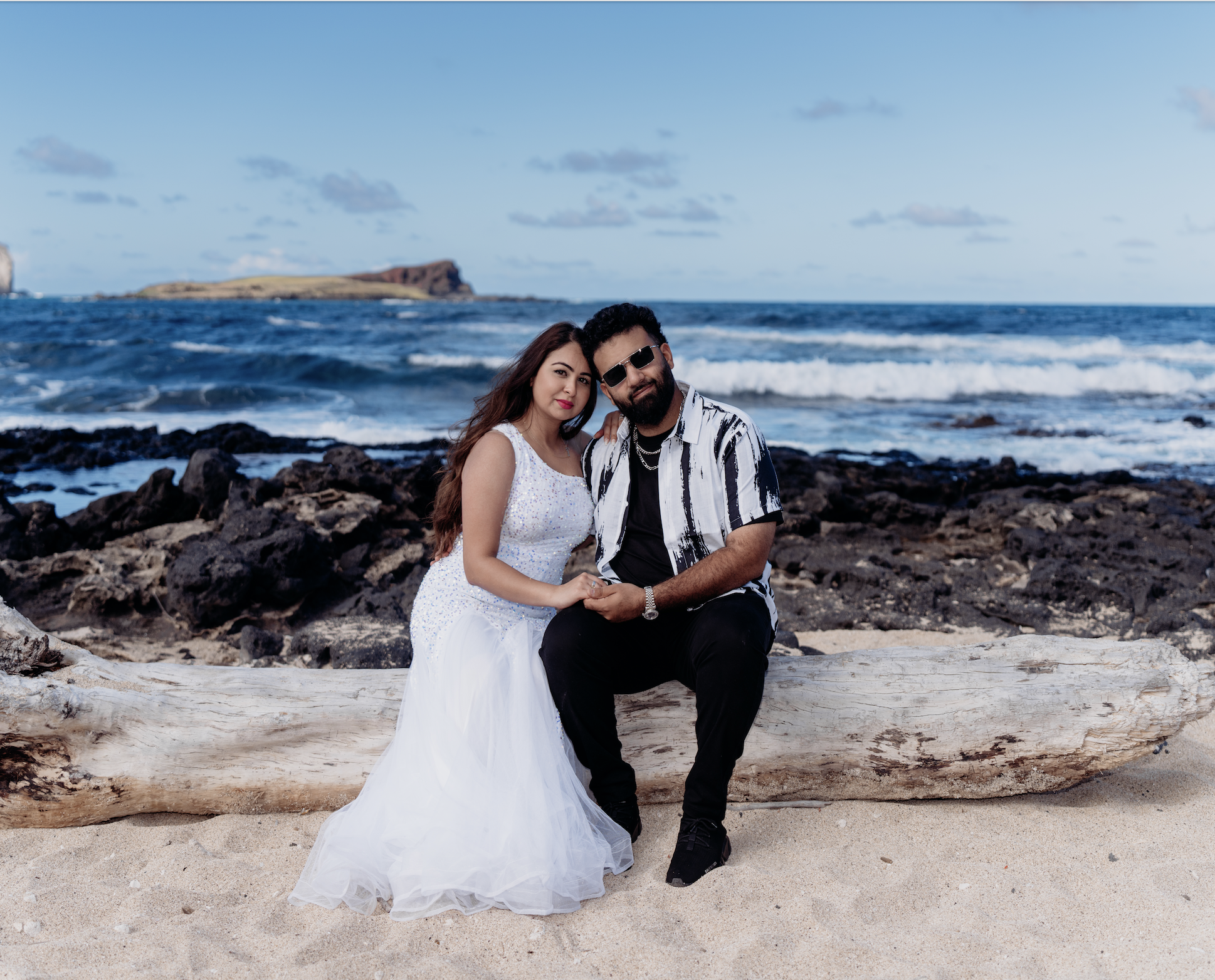 A couple sitting on a large piece of driftwood on a sandy beach, with the ocean and rocky shoreline in the background. The woman is wearing a white dress, and the man is dressed in a black and white striped shirt with black pants and sunglasses.