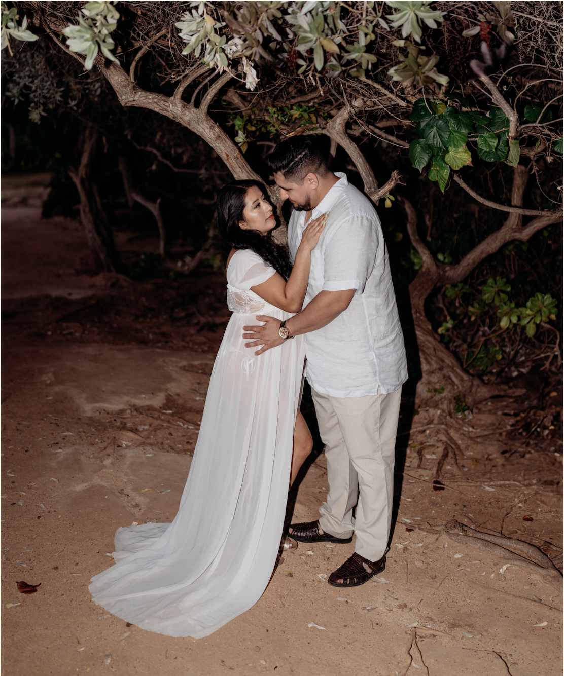 A couple standing close together on a dirt ground at night, under a tree with twisting branches and green leaves, gazing into each other's eyes. The woman is wearing a white dress with lace details, and the man is in a white shirt and beige pants.