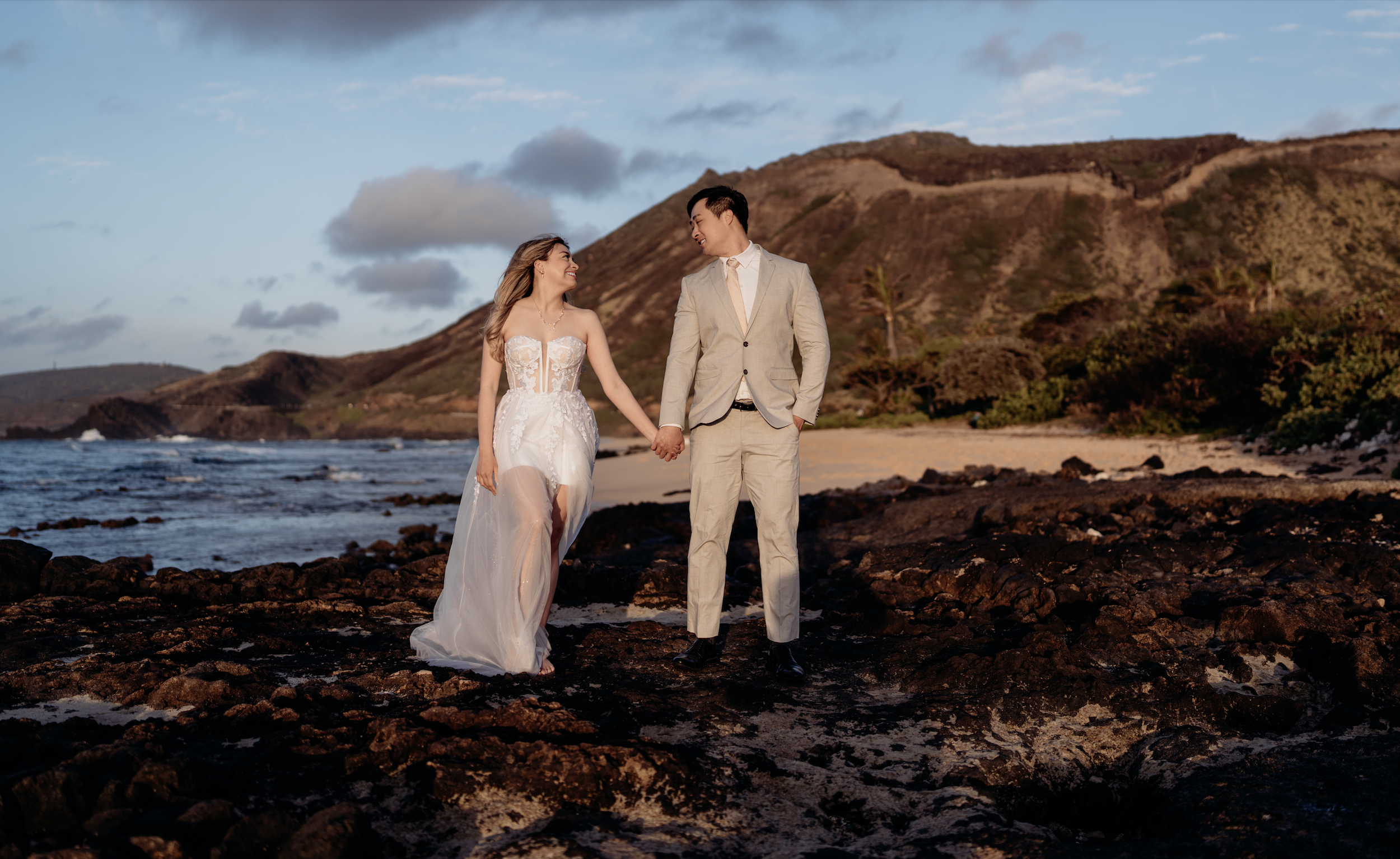A newlywed couple holding hands on the rocky beach during sunset, with the ocean, sandy shore, and hills with greenery in the background.