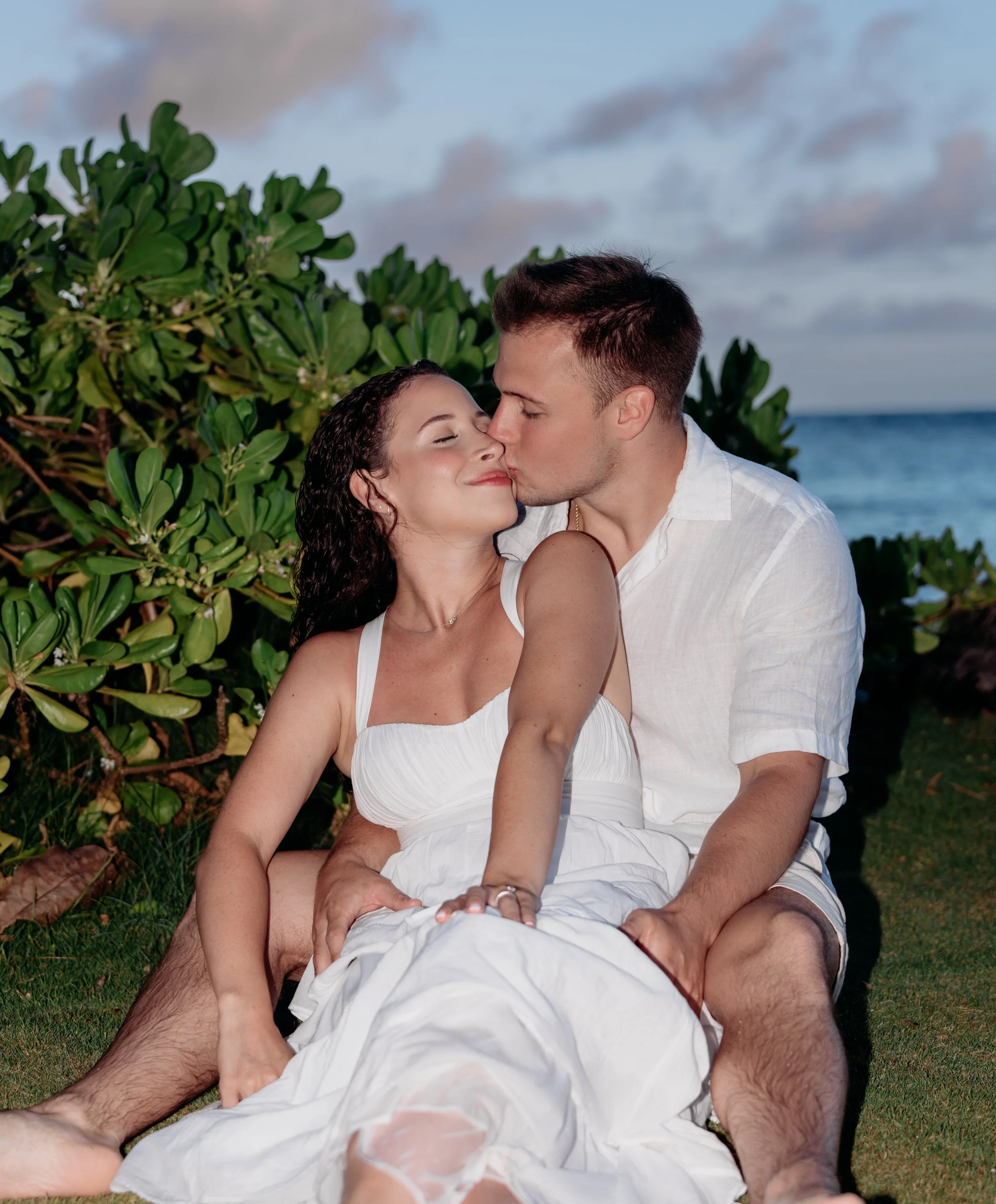 A couple sitting on grass near the beach, with greenery and ocean in the background, sharing a kiss and enjoying a romantic moment.