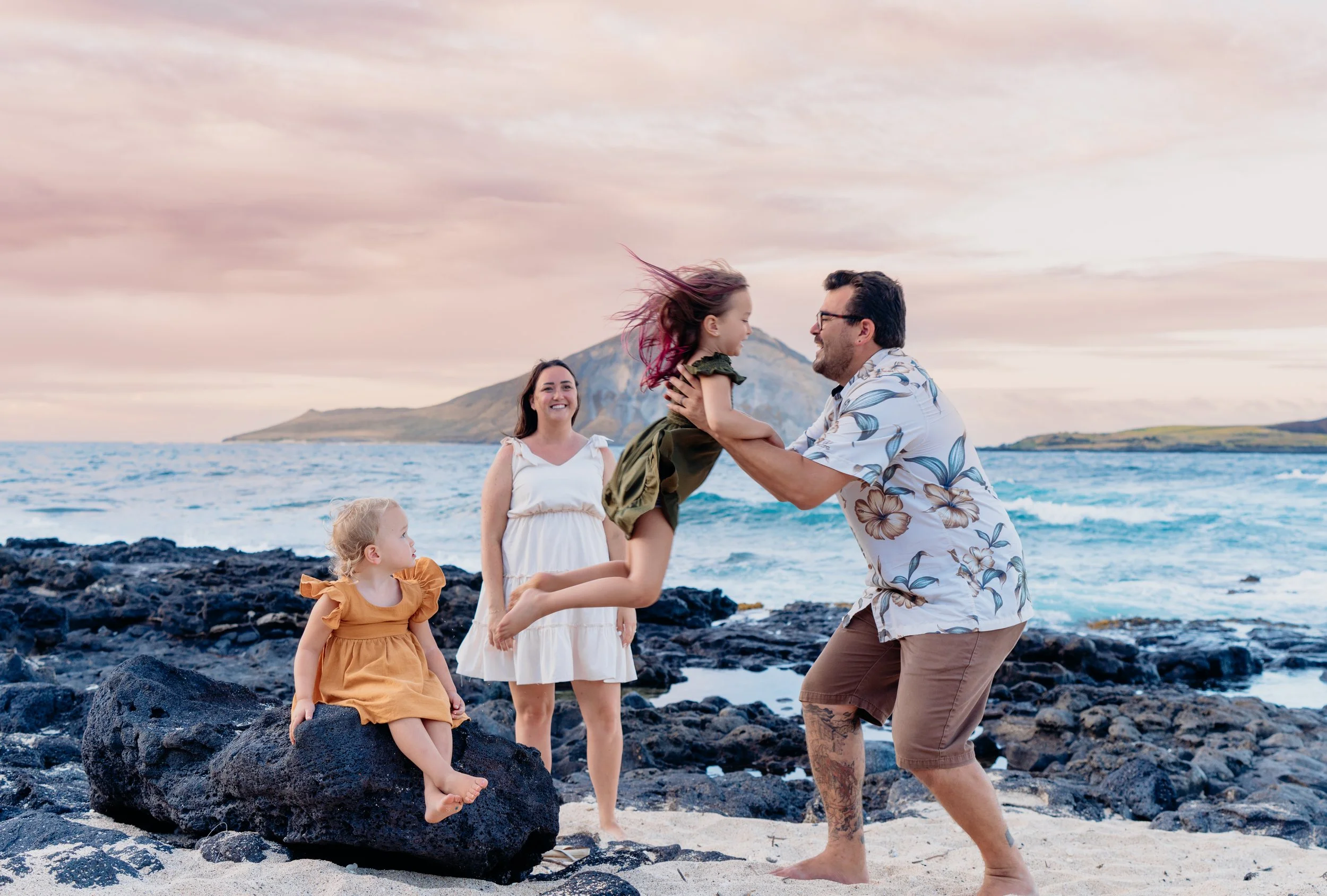 Family on a rocky beach enjoying sunset, with a man lifting a girl in a green dress, women and a younger girl watching, ocean in the background.