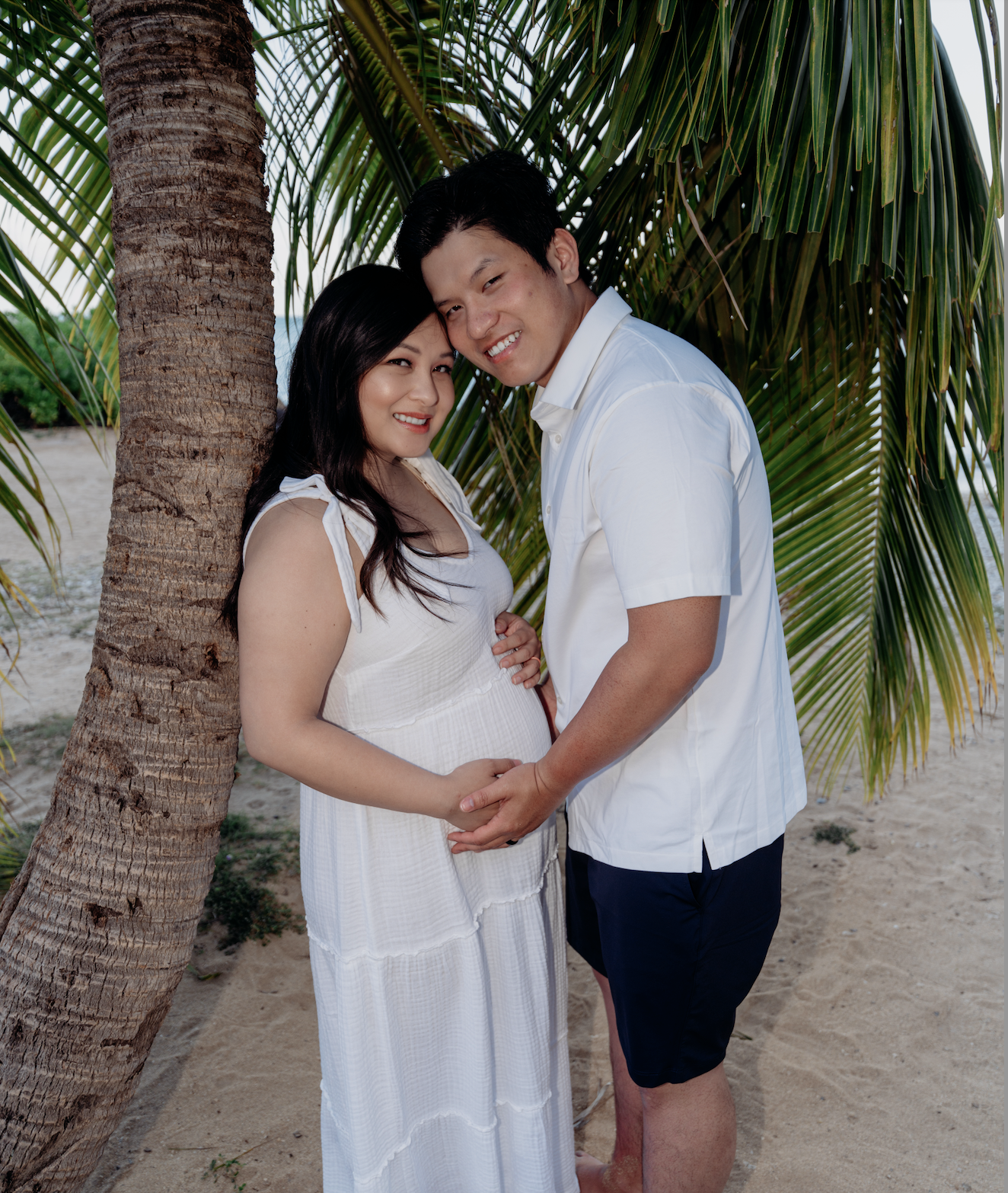 A couple standing on a sandy beach, hugging each other and smiling. There are palm trees in the background, and they are dressed in white summer clothing.