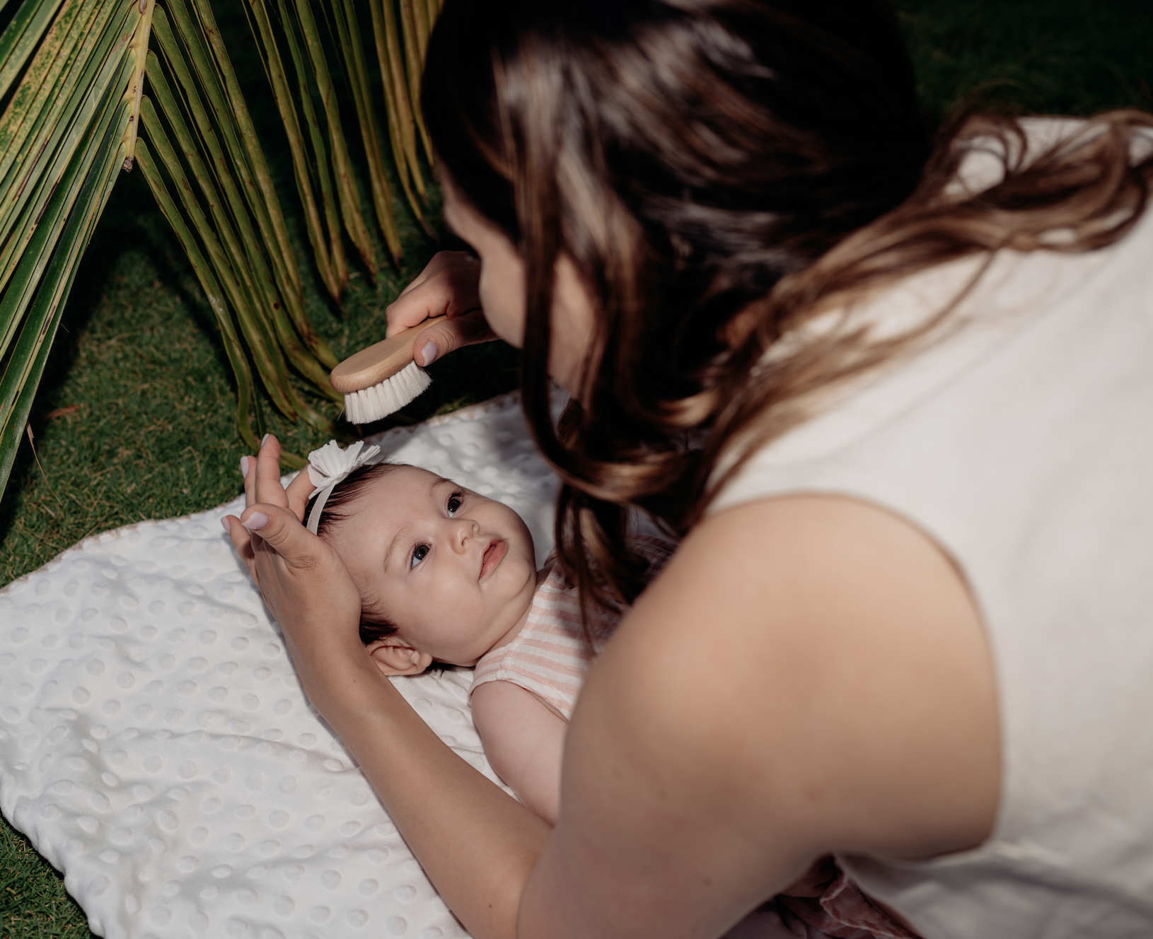 A woman is grooming a baby girl with a brush while lying on a white blanket outdoors near green grass and plants.