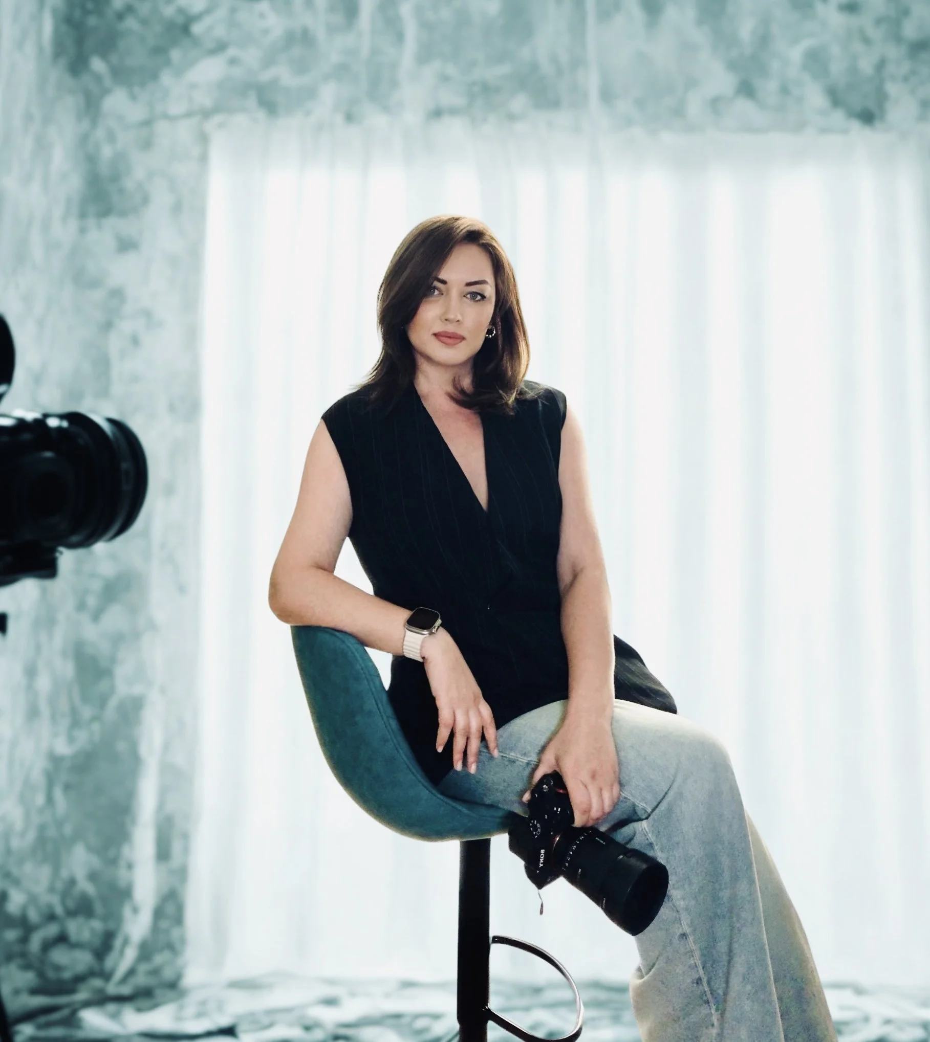 A woman sitting on a teal chair in front of a bright window with sheer curtains, with a camera pointed at her, wearing a black sleeveless top, gray pants, and a smartwatch.