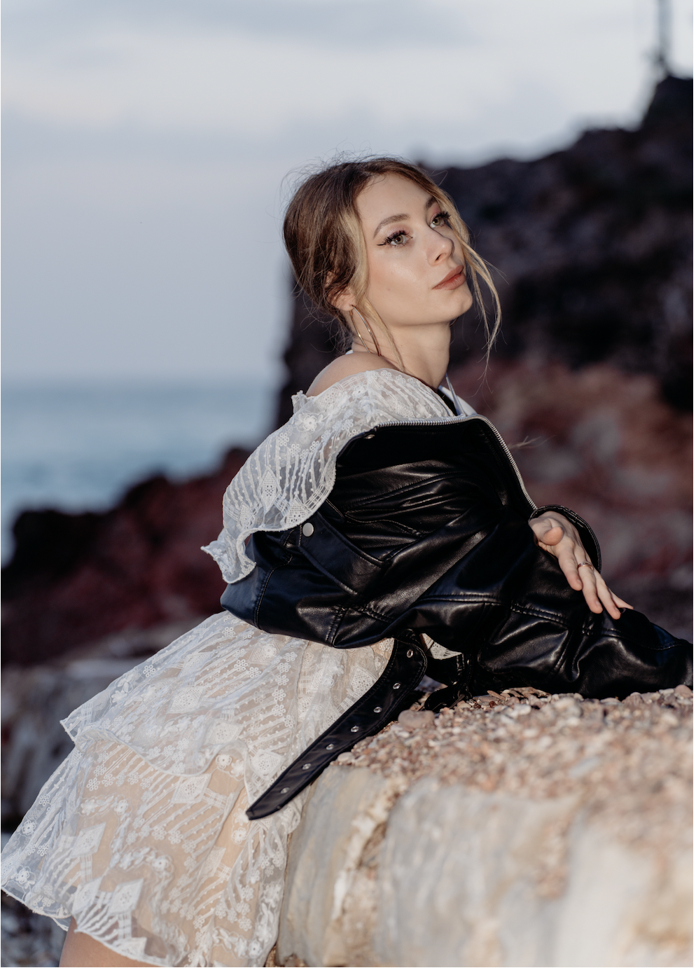 A young woman with light brown hair, wearing a white lace dress and a black leather jacket, leaning on a rocky surface near the beach during sunset.