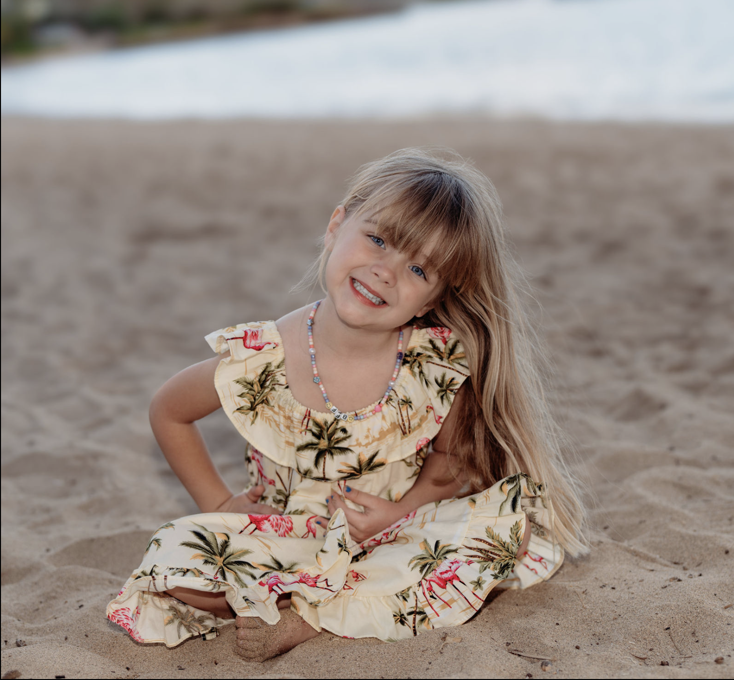 A young girl in a floral dress sitting on sandy beach with ocean waves in the background, smiling cheerfully.