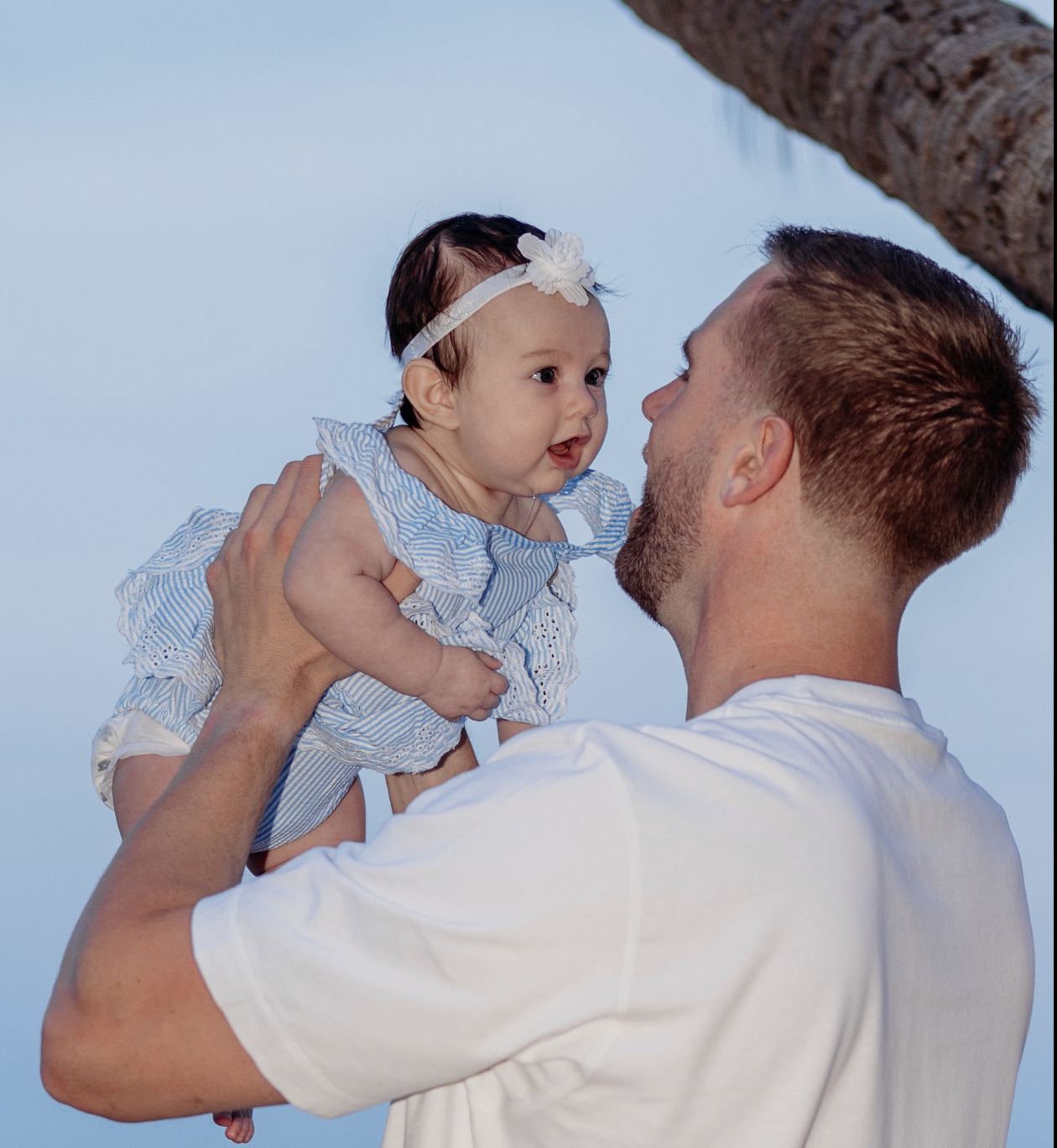 A man holding a smiling baby girl wearing a blue and white striped dress and a headband with a flower, outdoors near a tree with a blue sky background.