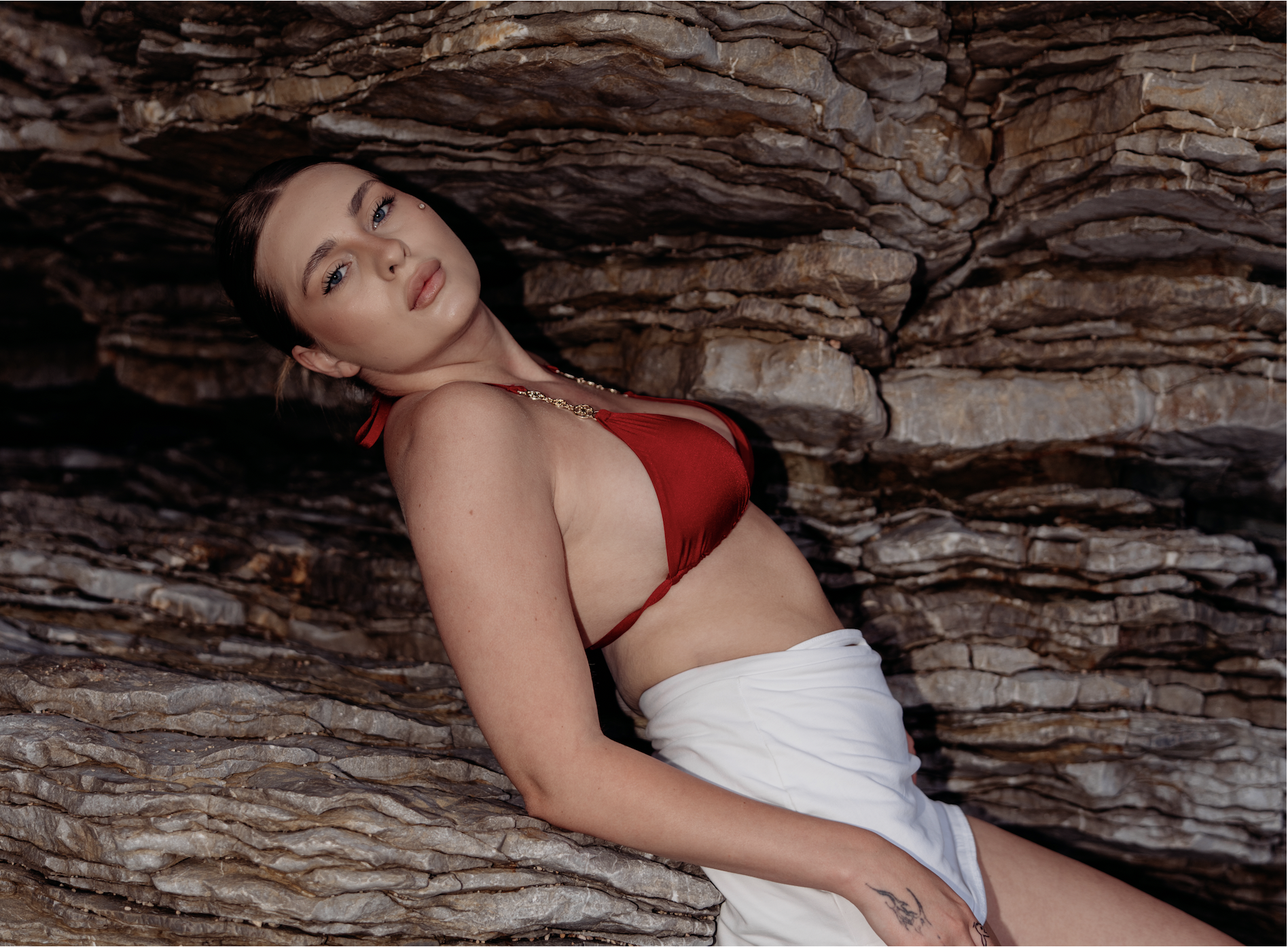 Young woman with dark hair and blue eyes leaning against a textured rock wall, wearing a red bikini top, a gold necklace, and white bottoms.