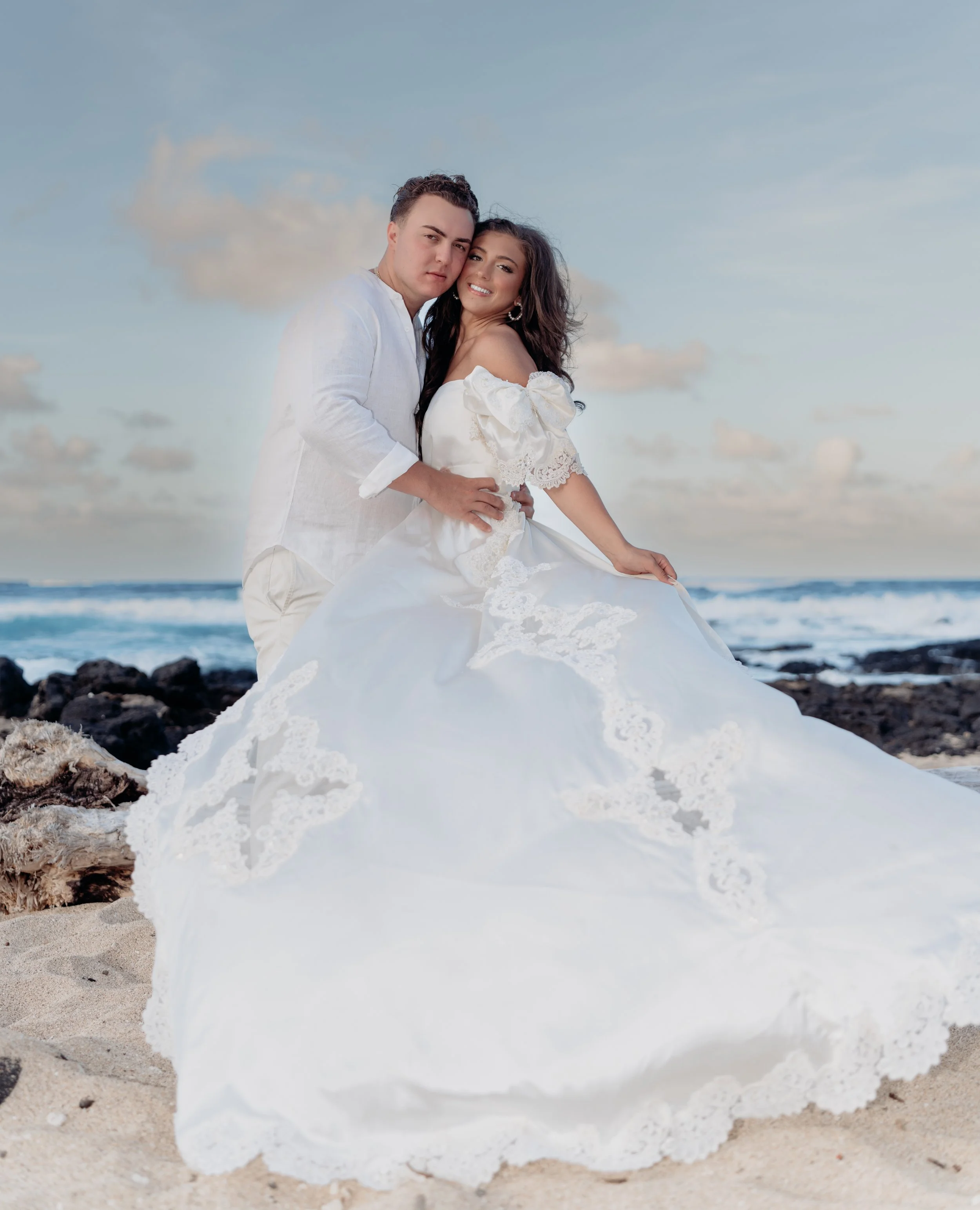 A couple in wedding attire on a beach with rocks and ocean waves, the groom in a white shirt and the bride in a white dress with lace, posing together under a partly cloudy sky.