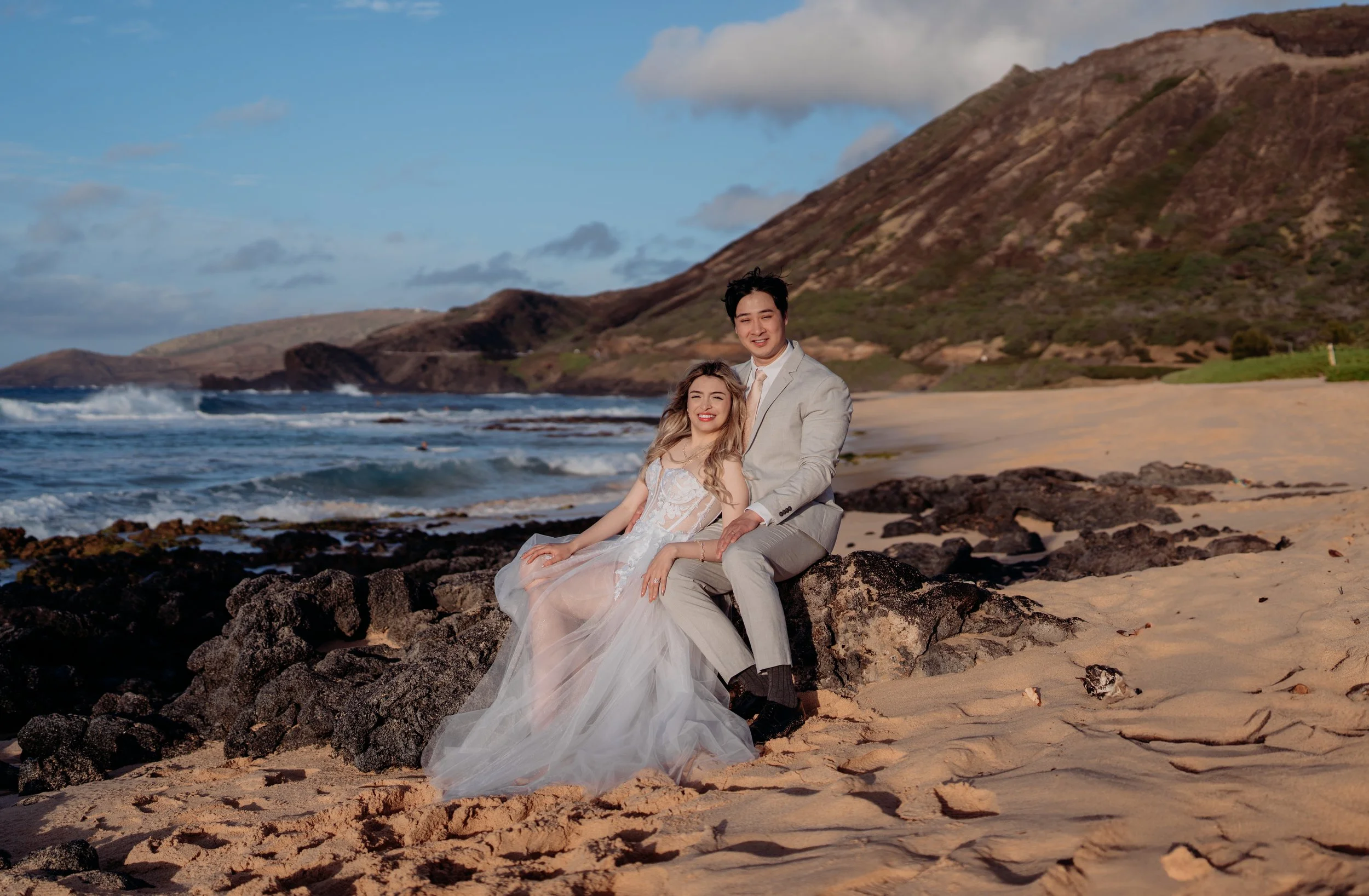 A bride and groom in wedding attire sitting on rocks at a beach with sand, ocean waves, and hills in the background during sunset.