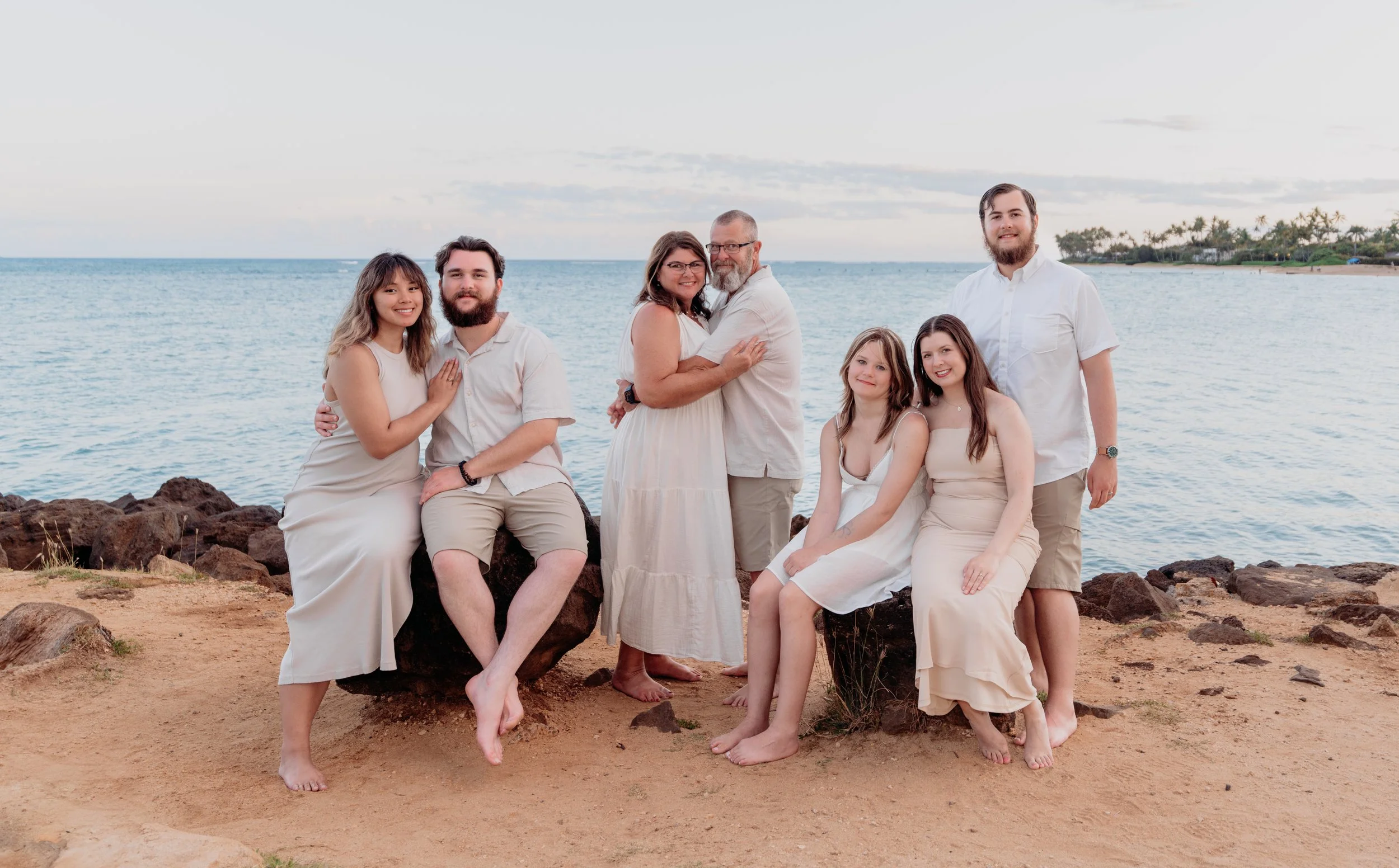 Family of seven dressed in light-colored clothes at the beach, sitting and standing on sandy ground with rocks, ocean behind them, during sunset.