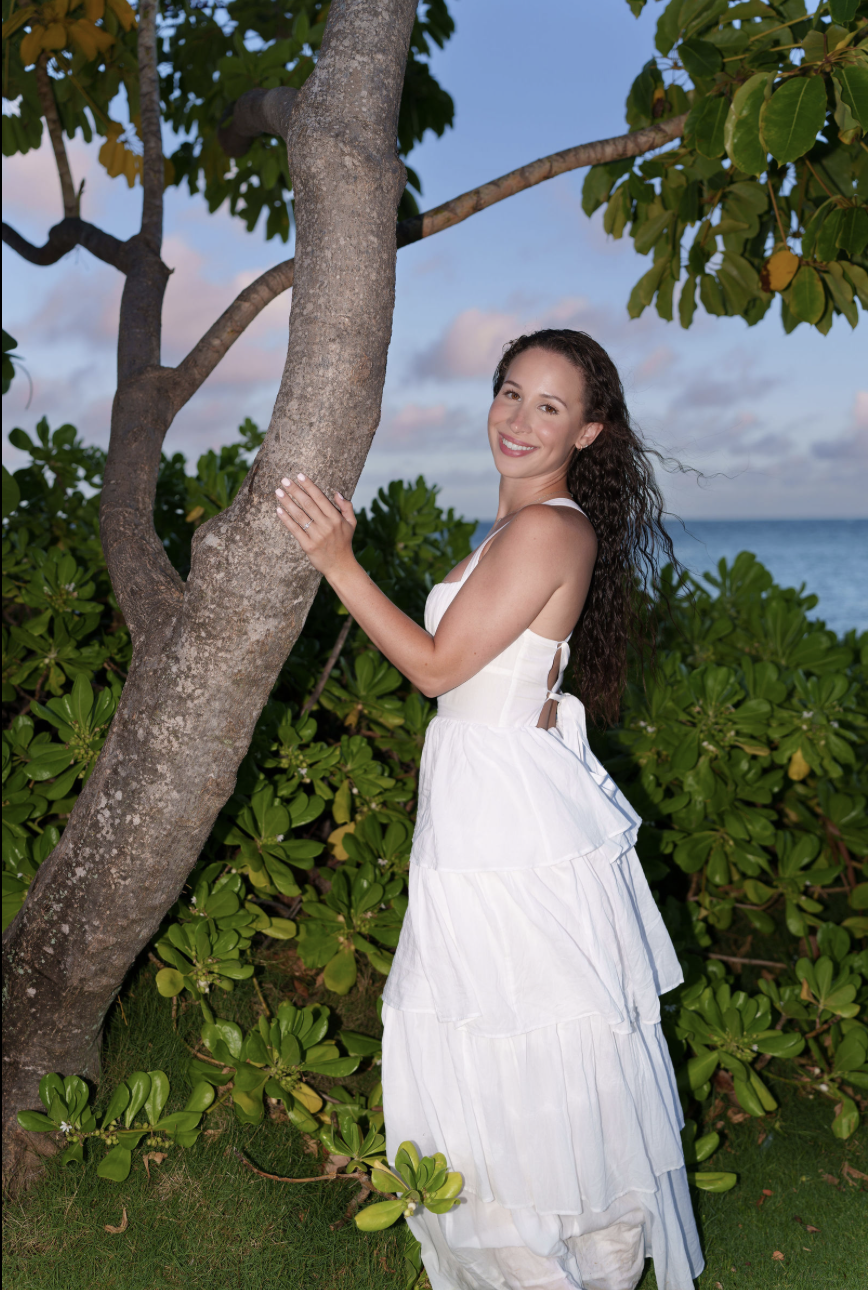 A smiling woman with long, curly brown hair wearing a white sleeveless dress standing beside a tree with green leaves, with an ocean and a partly cloudy sky in the background during daytime.