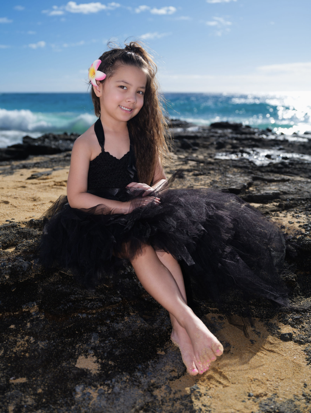 Young girl in a black dress with a pink and yellow flower in her hair sitting on rocks by the beach, smiling with the ocean and blue sky in the background.