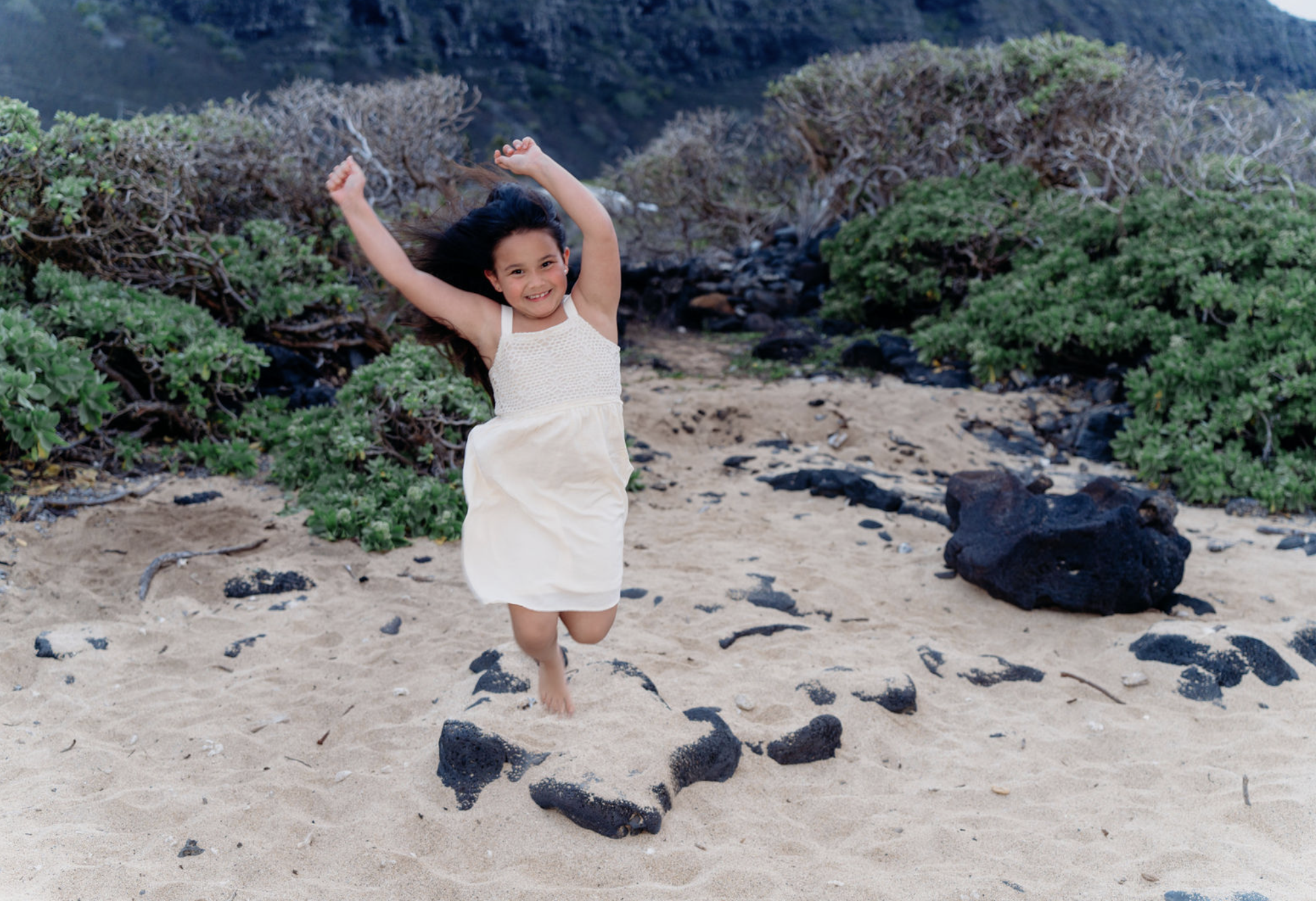 A young girl in a cream-colored dress jumping on a sandy beach with volcanic rocks and green bushes in the background.