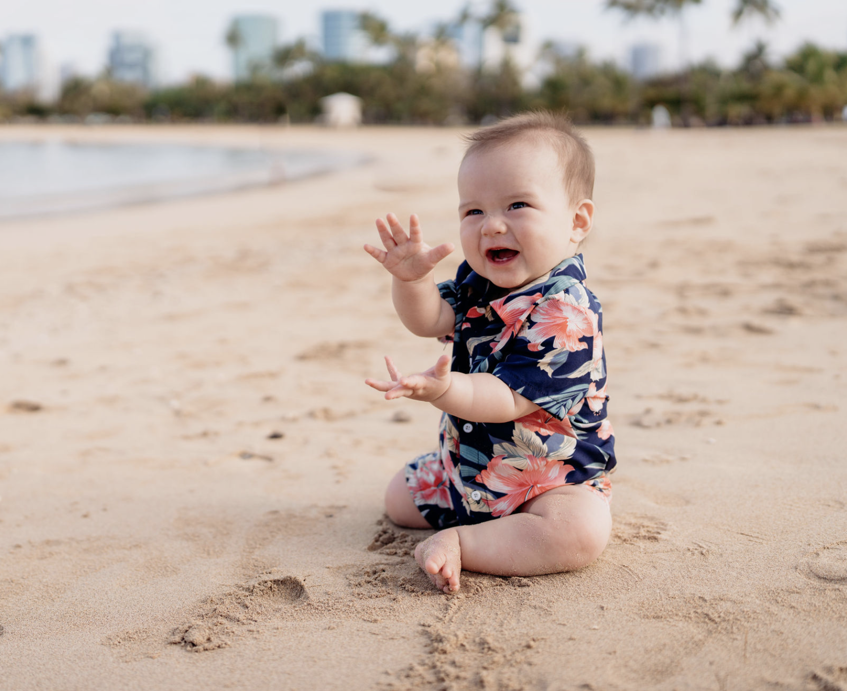 A baby sitting on a sandy beach wearing a colorful Hawaiian shirt, smiling and waving with one hand, background of the ocean and city skyline.