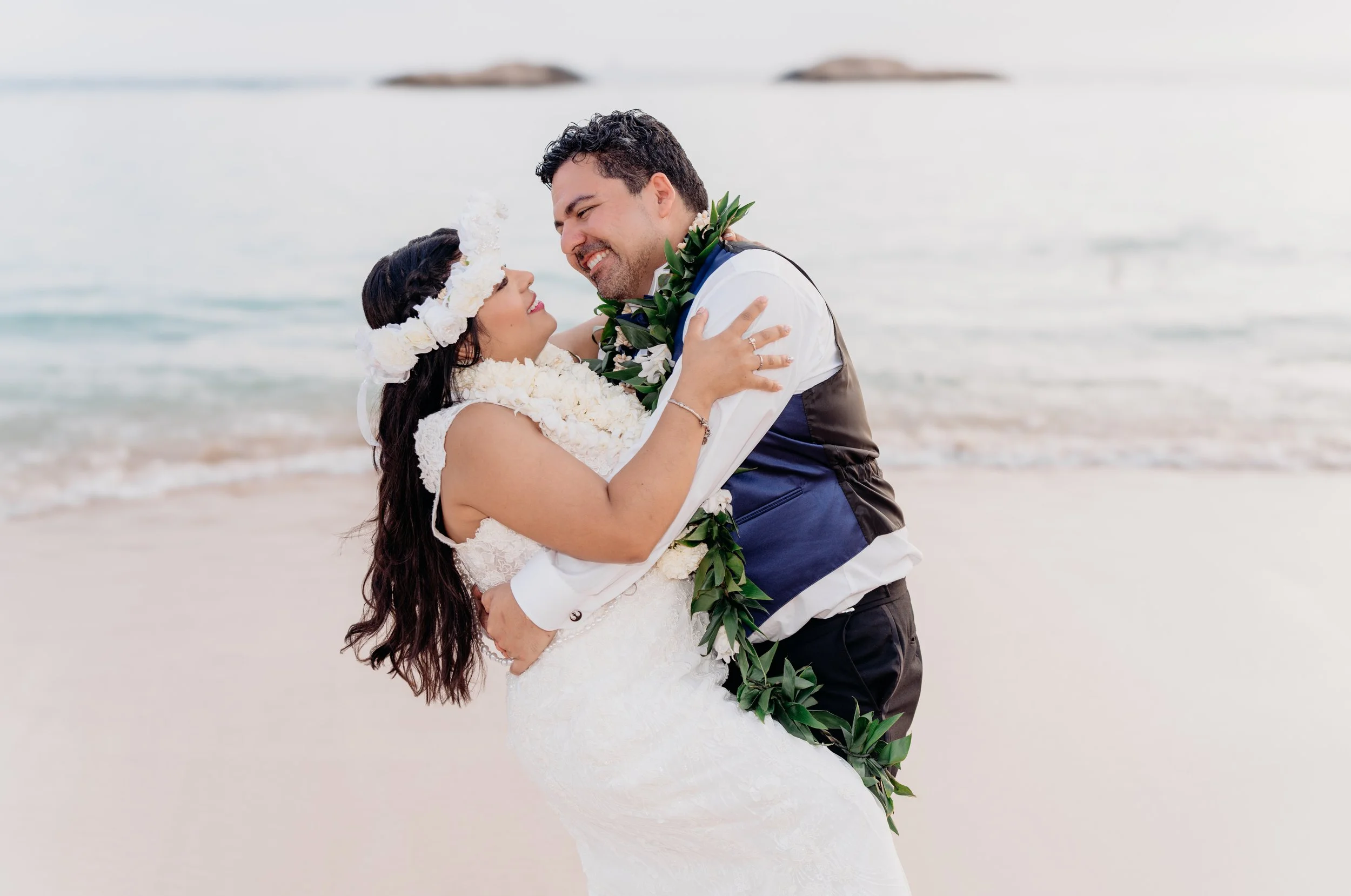 A newlywed couple dressed in wedding attire on a beach, embracing and smiling at each other, with the ocean and rocks in the background.