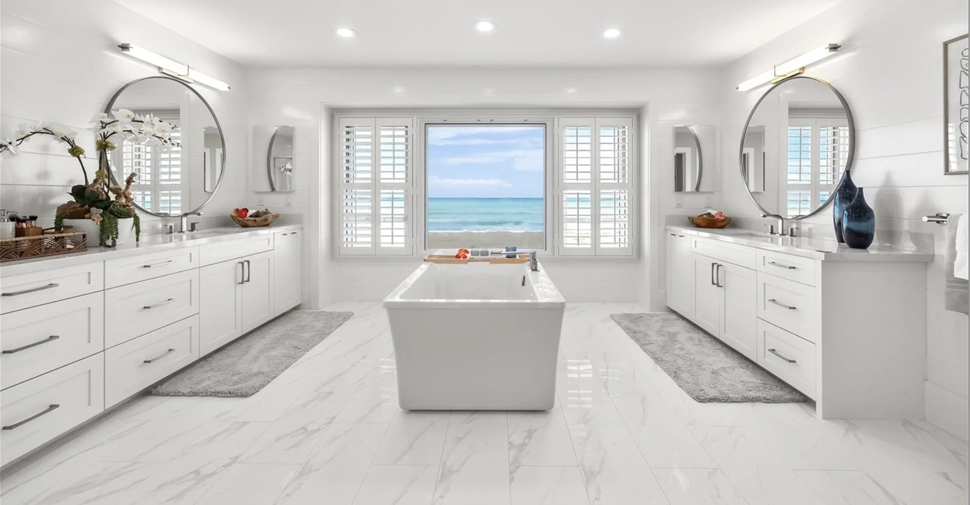 Bright bathroom with white cabinets, a freestanding bathtub, large window with ocean view, and decorative plants and vases.
