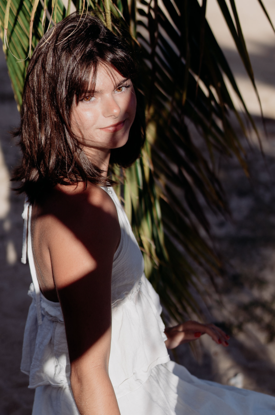 A woman with shoulder-length brown hair and light skin sits outdoors near a palm tree, wearing a white dress, with sunlight casting shadows on her face and body.