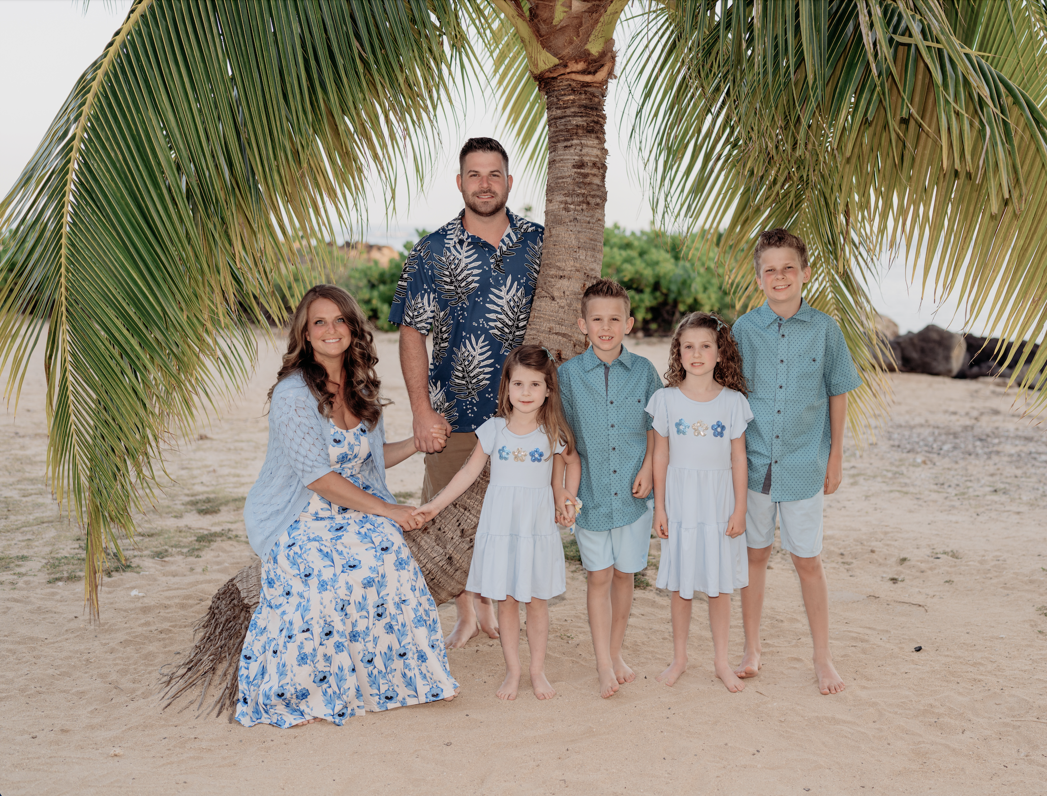 A family of seven posing on a sandy beach under a palm tree, with the ocean and rocks in the background. The mother, father, three boys, and two girls are dressed in blue and white summer clothes. The mother is seated on the sand, and the others stan