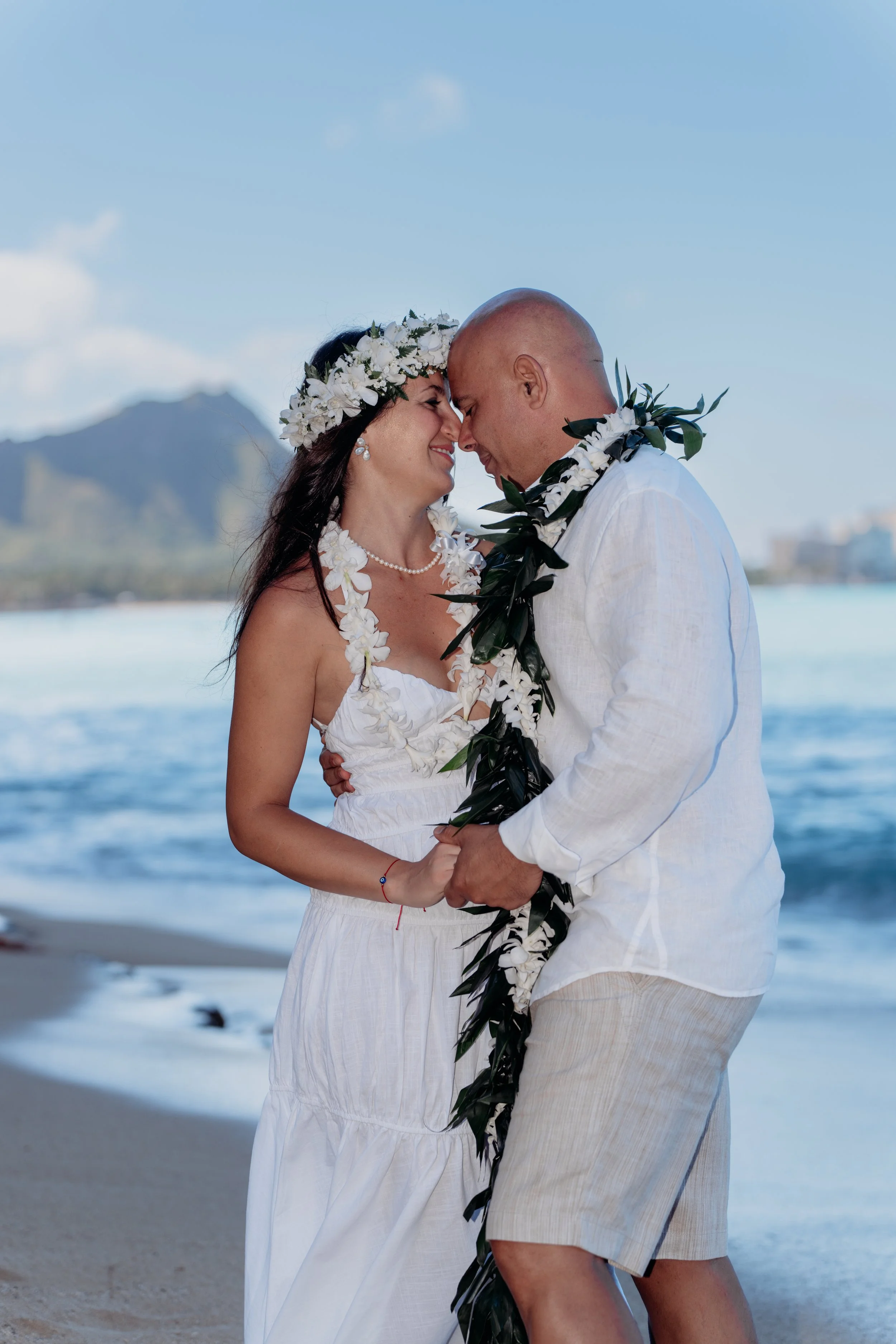 A couple dressed in wedding attire holding hands on a beach, with a mountain and ocean in the background, during daytime.