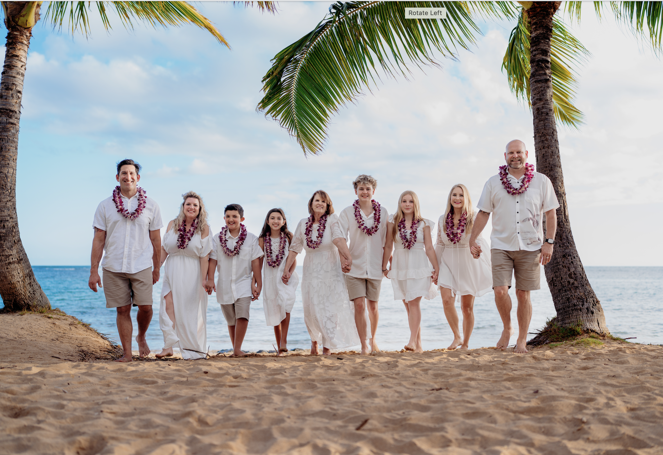 A family of nine, dressed in white, walking barefoot on the beach under palm trees during daytime, wearing flower leis, and holding hands.